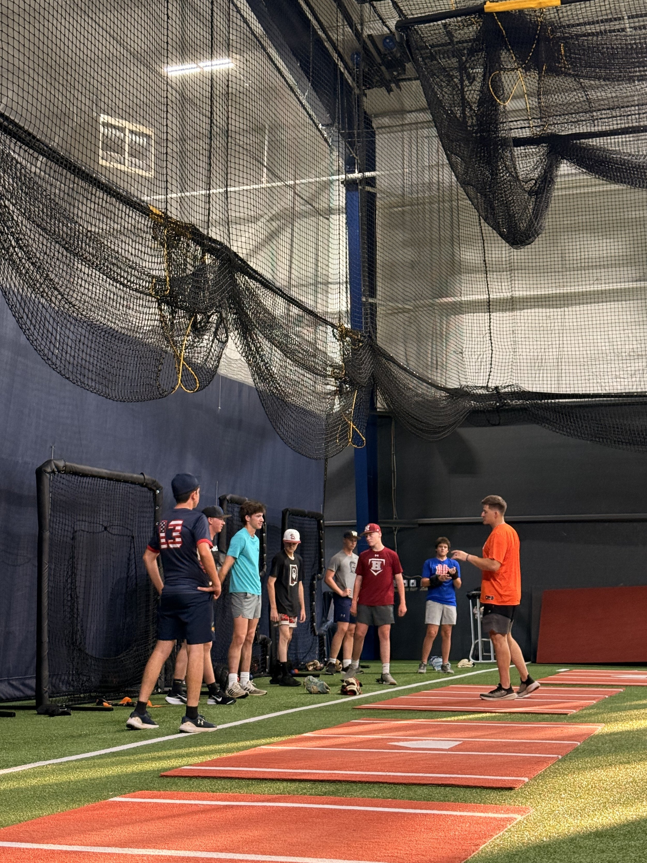 Group of young athletes standing on a turf field next to orange agility mats, with an indoor sports facility in the background, and one coach or trainer talking to them.