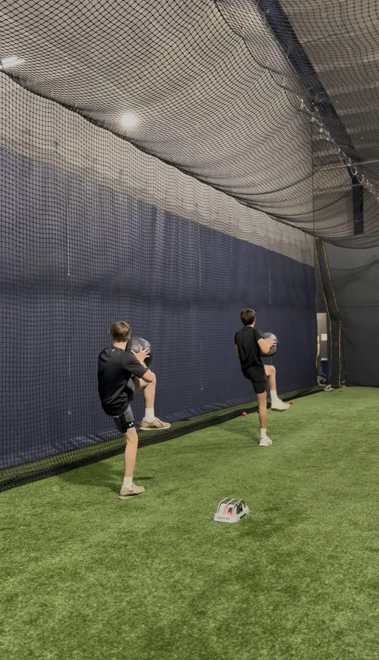 Three boys in black athletic clothing holding medicine balls during a workout inside a gym with artificial turf and a netted wall.