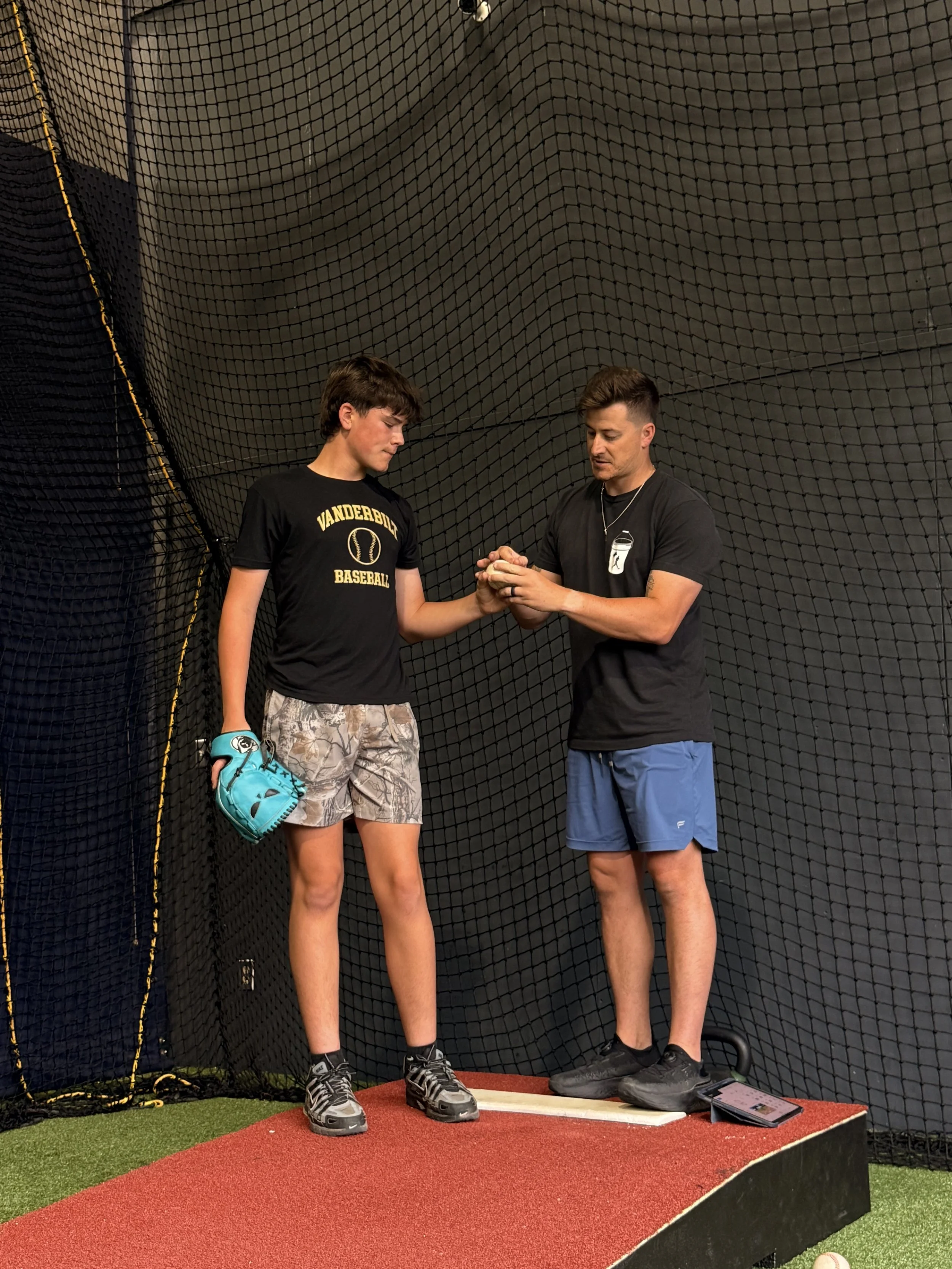 Two young men standing on a platform with a baseball net background. The one on the left holds a baseball glove and is wearing a "Vanderbilt Baseball" t-shirt, camouflage shorts, and sneakers. The man on the right is holding the other's hand, wearing a black t-shirt with a graphic, blue shorts, and black shoes, and is possibly coaching or instructing the other.
