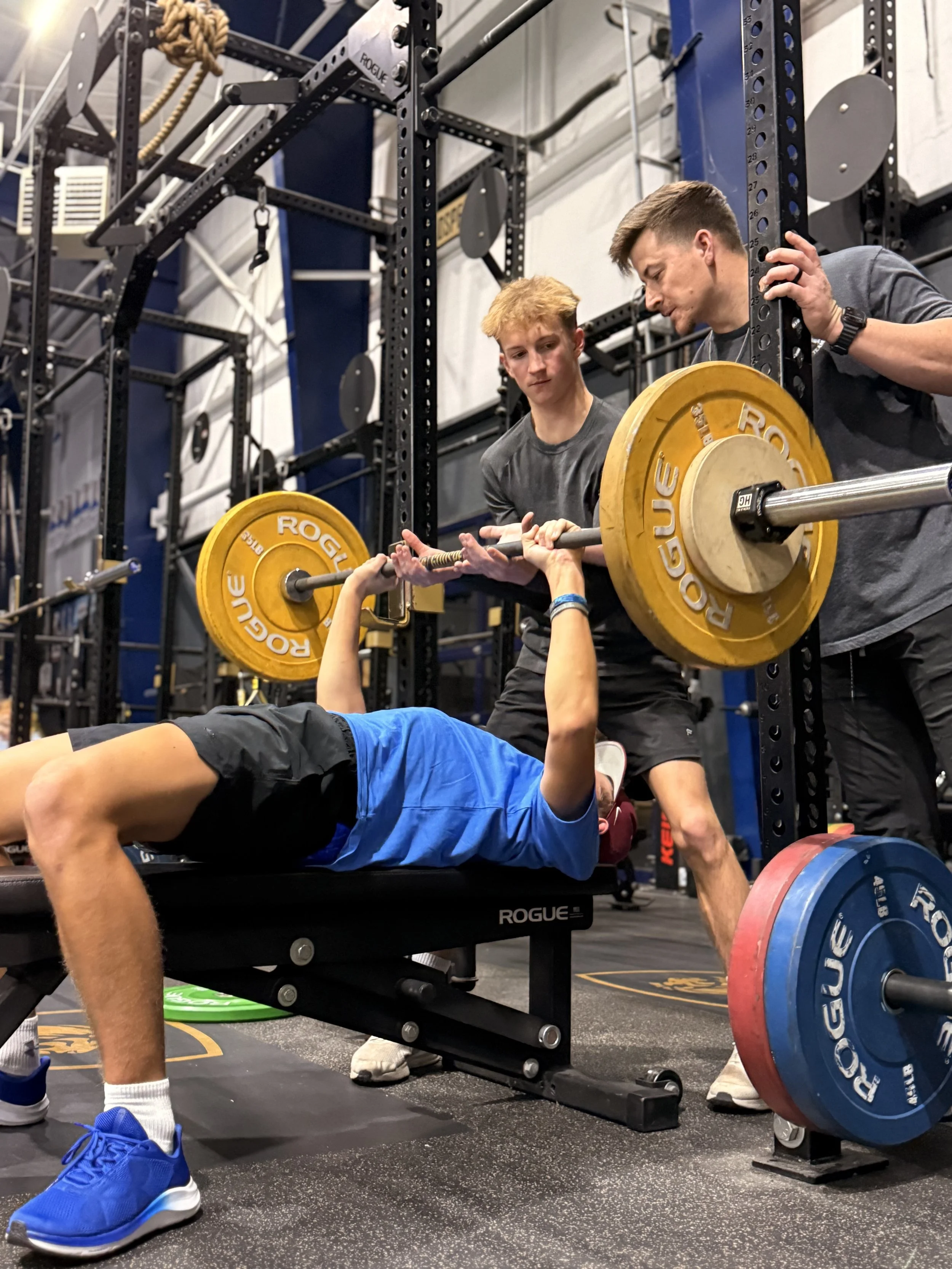 Two young men in a gym helping a person who is lying on a bench press to lift a barbell with yellow and blue weight plates. The gym has metal racks, ropes, and equipment in the background.