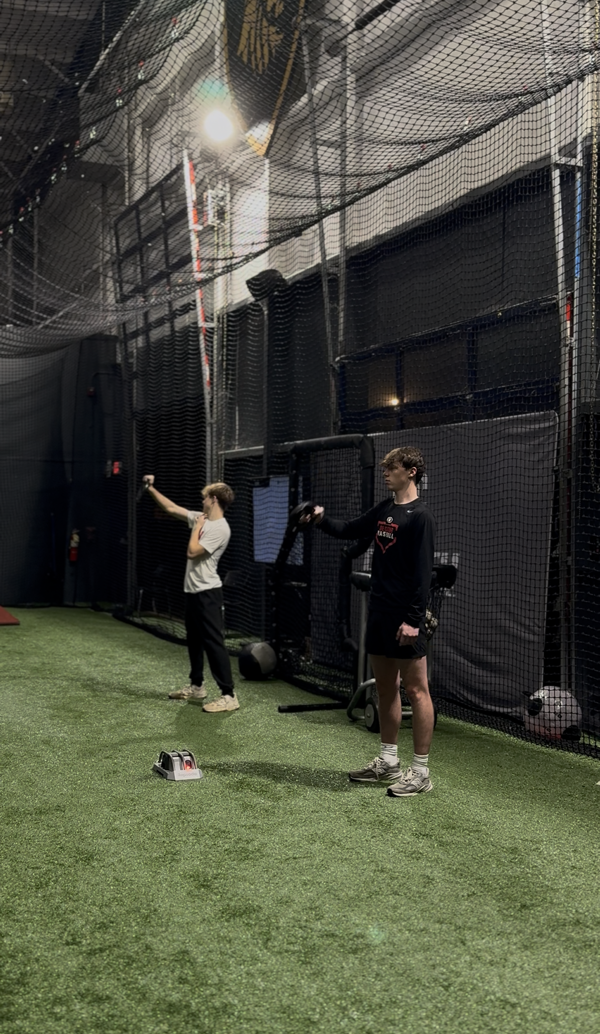 Two young men in athletic clothing doing exercises with resistance bands in an indoor sports facility with artificial turf and black netting.