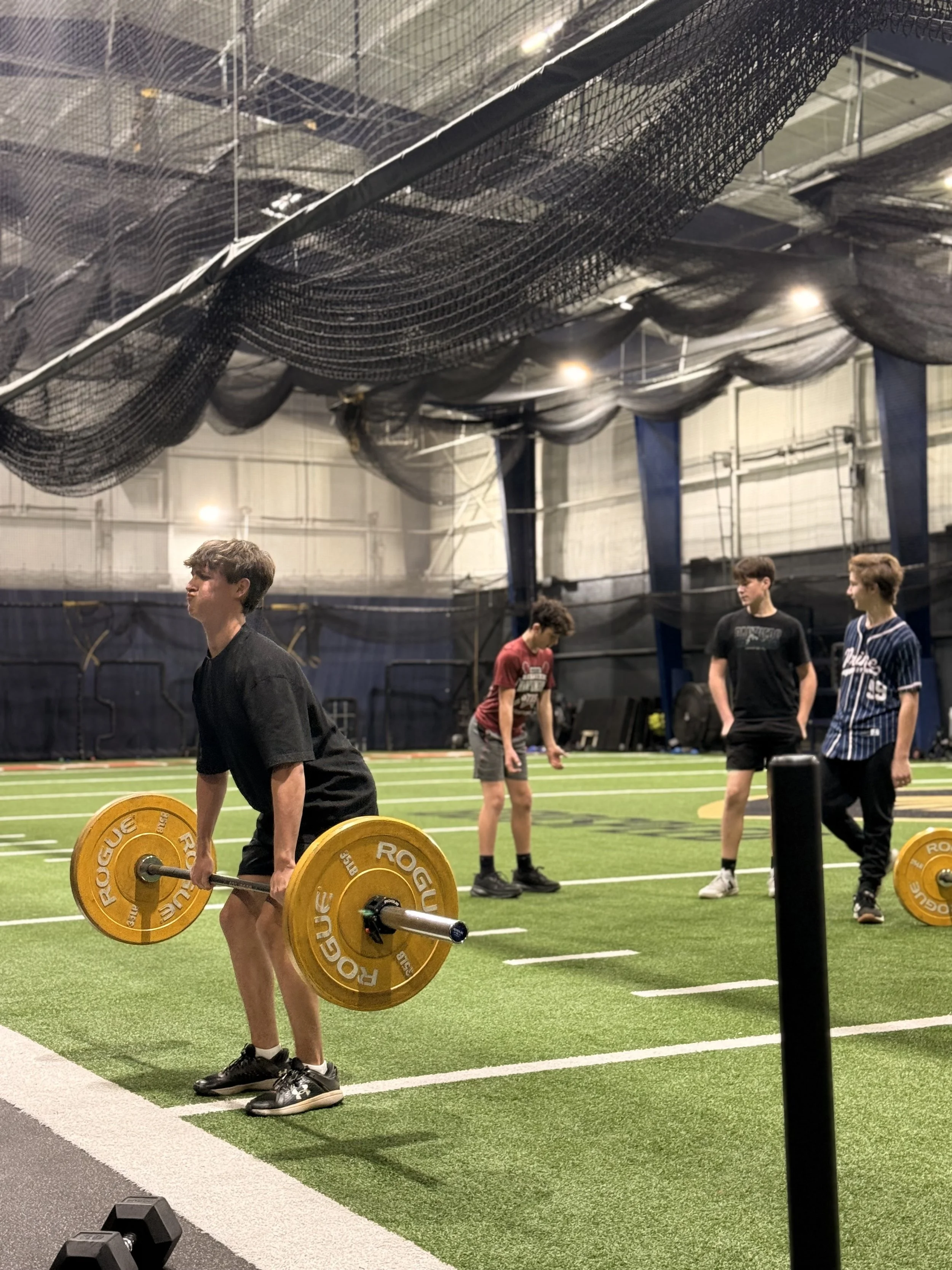 A young man lifting a barbell with orange weights at an indoor sports facility with other young people in the background.