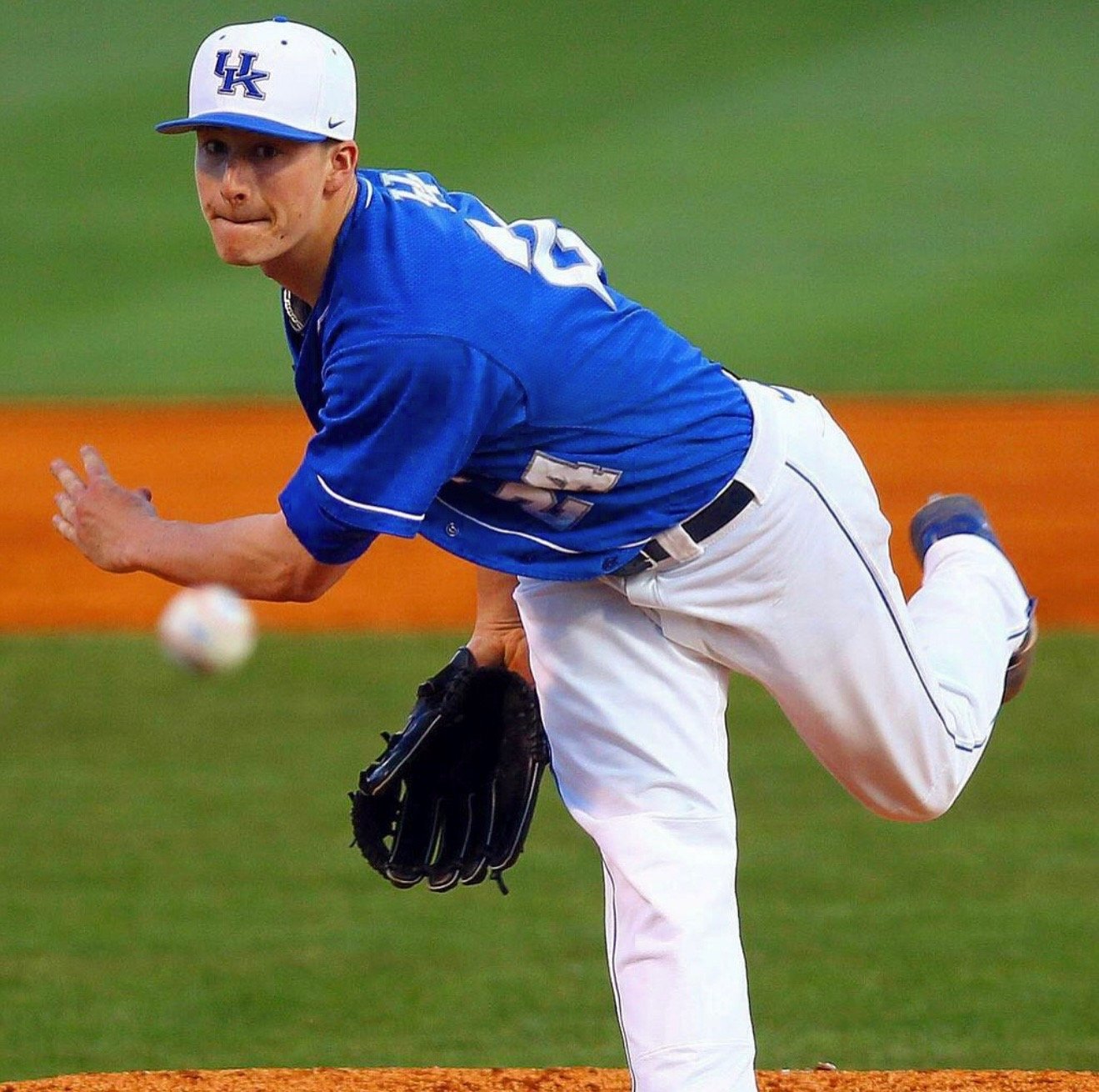 A baseball player wearing a blue jersey with 'UK' on the cap, white pants, and a black glove, is pitching on the field.