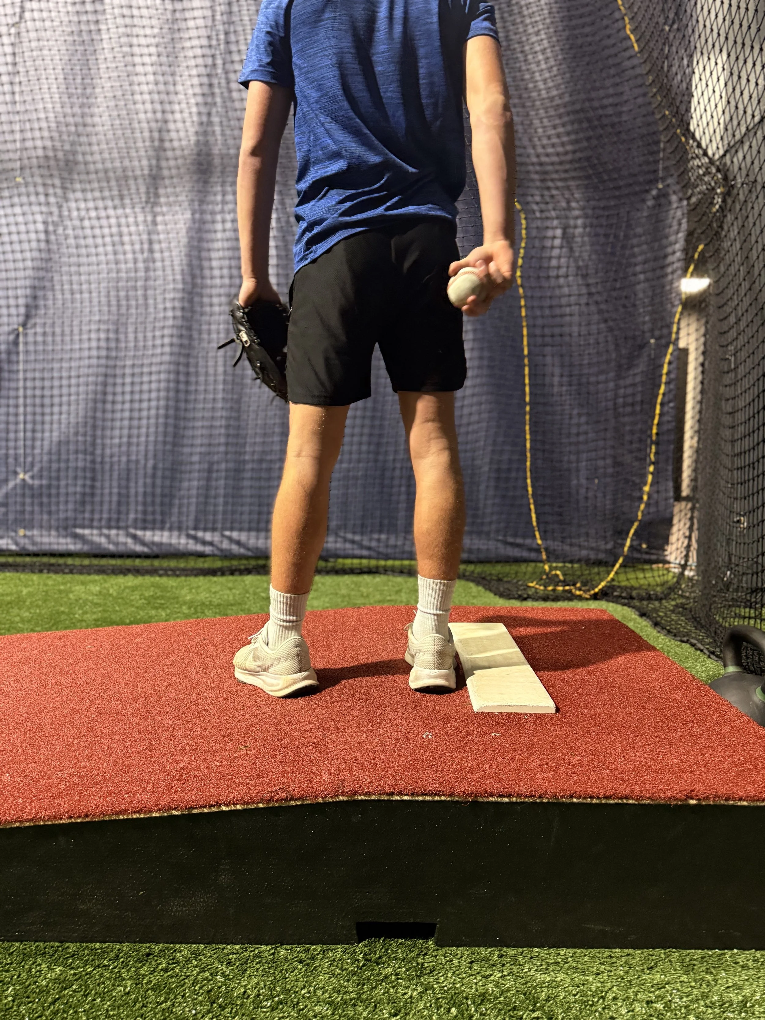 Person in athletic clothes holding a baseball and glove at a training net, preparing to throw or catch, standing on a red mat.