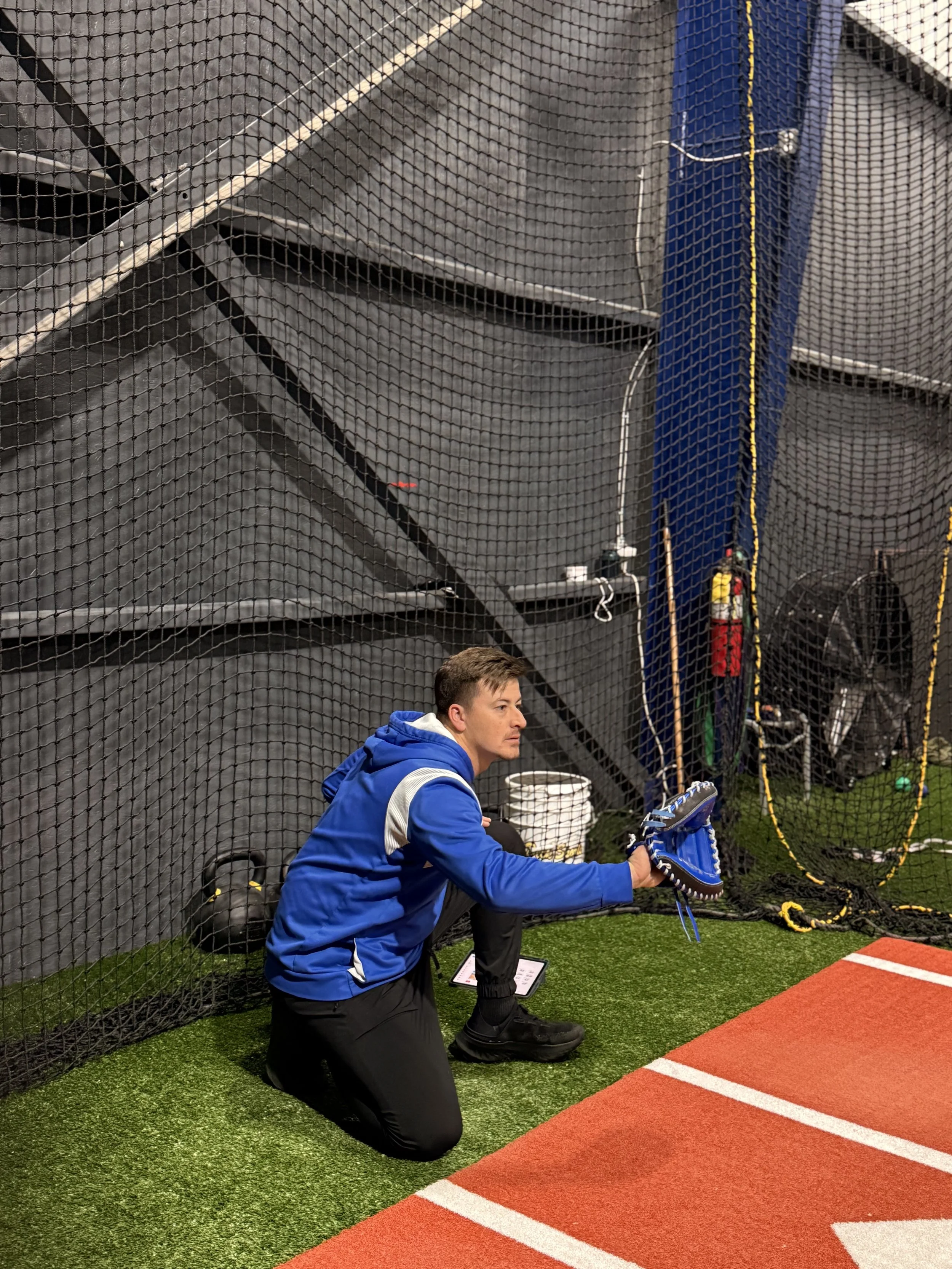 A man kneeling on an indoor sports field, holding a baseball glove, with a net and sports equipment nearby.