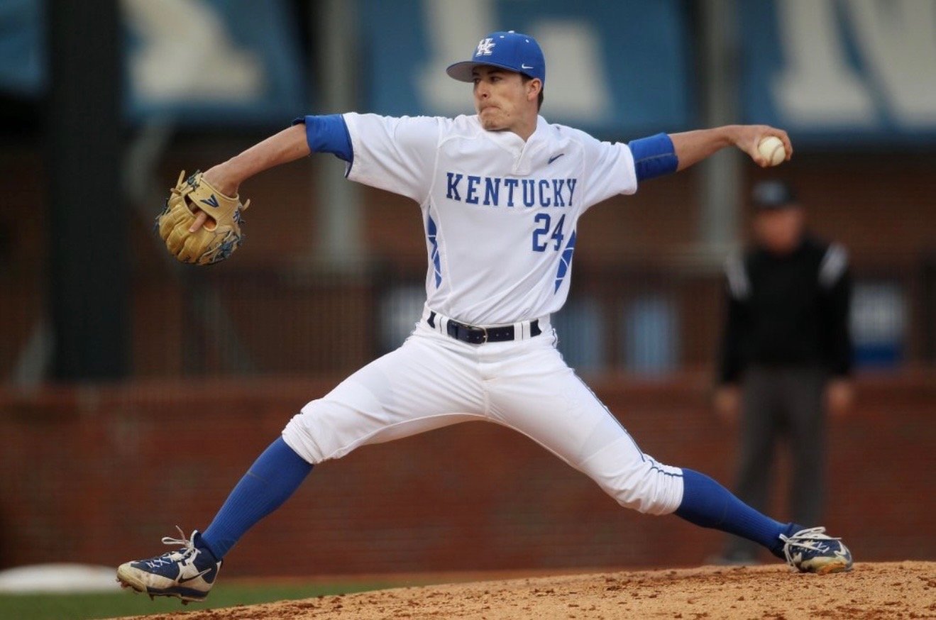 A baseball pitcher in a white Kentucky uniform with the number 24, wearing a blue cap, pitching on the mound with his right arm extended and holding a baseball, with a blurred background of a person watching and stadium structures.