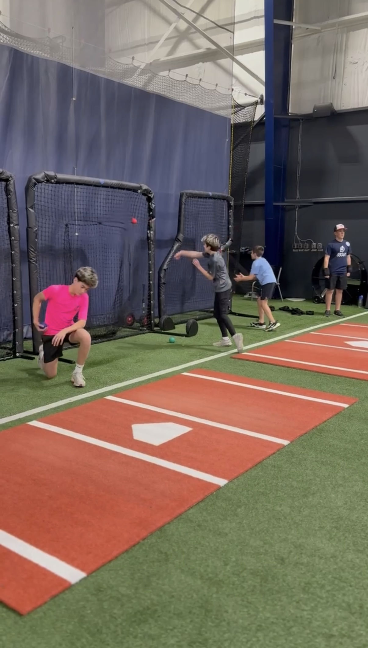 Children practicing baseball batting in an indoor sports facility with artificial turf, batting cages, and measured batting lanes.