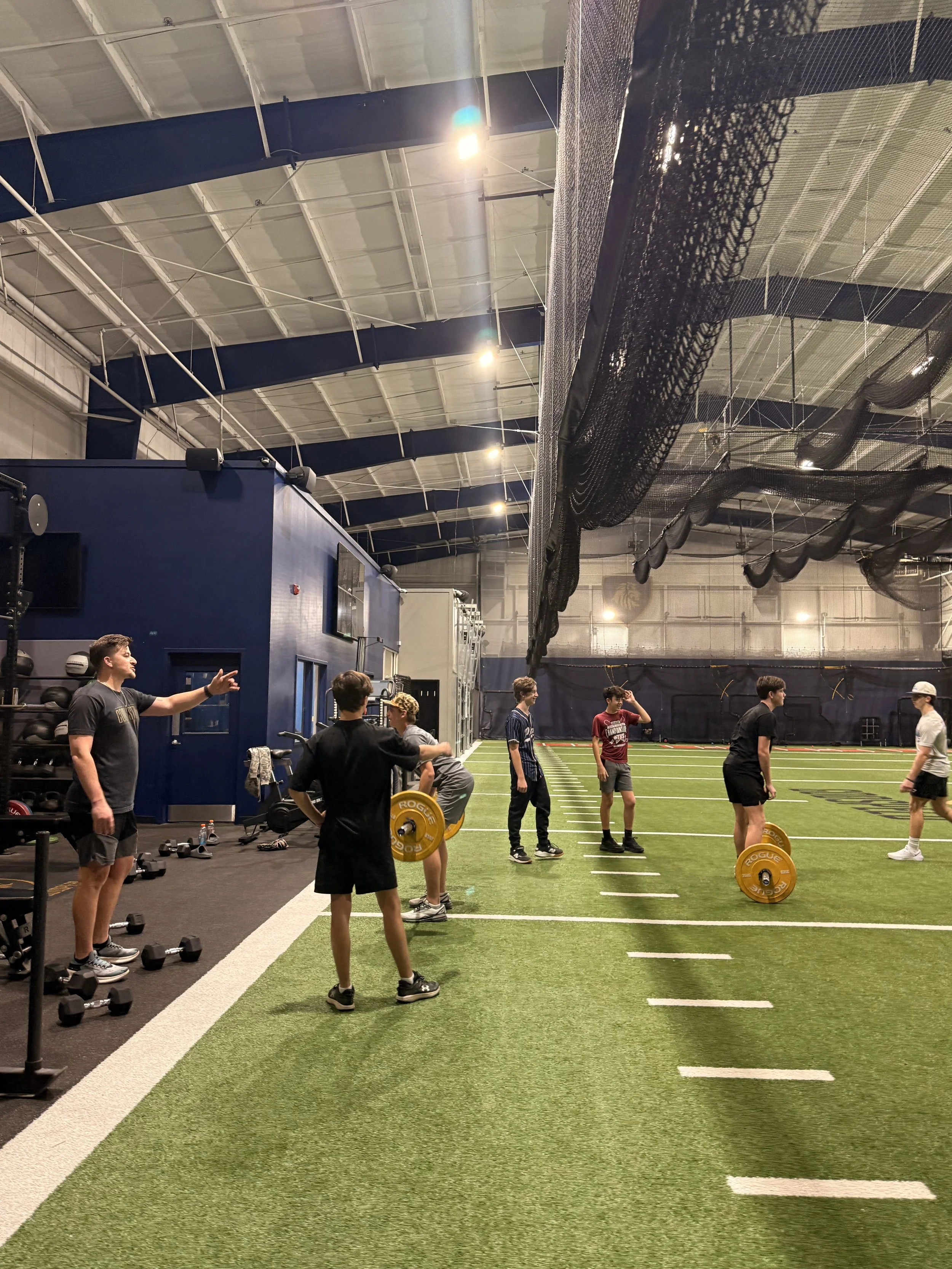Indoor gym with artificial turf, weightlifting equipment, and several young men participating in a workout session, under industrial ceiling with black nets hanging.