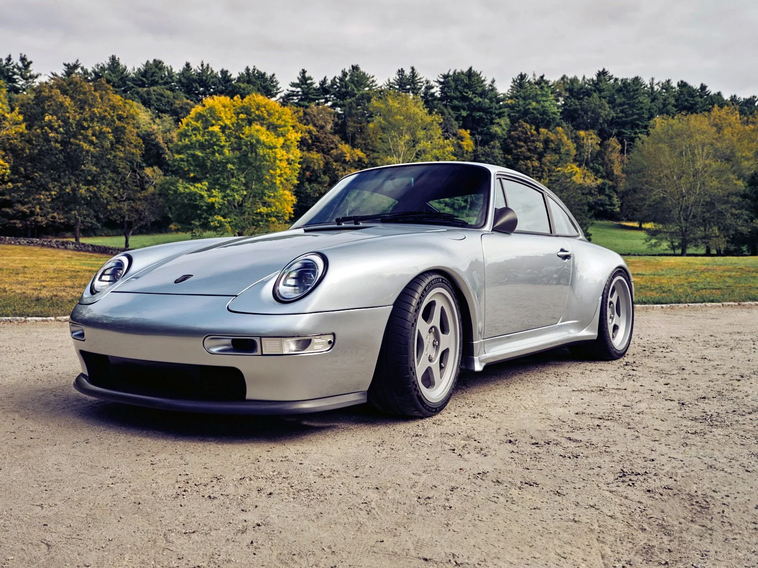 A silver Porsche 911 sports car parked on a gravel surface with trees and a grassy area in the background.