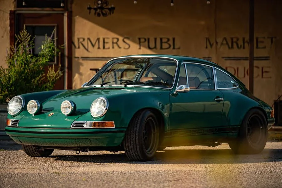 Green vintage Porsche 911 with four auxiliary lights on the front, parked in front of a rustic building with signs reading "FARMERS PUBL" and "MARKET".
