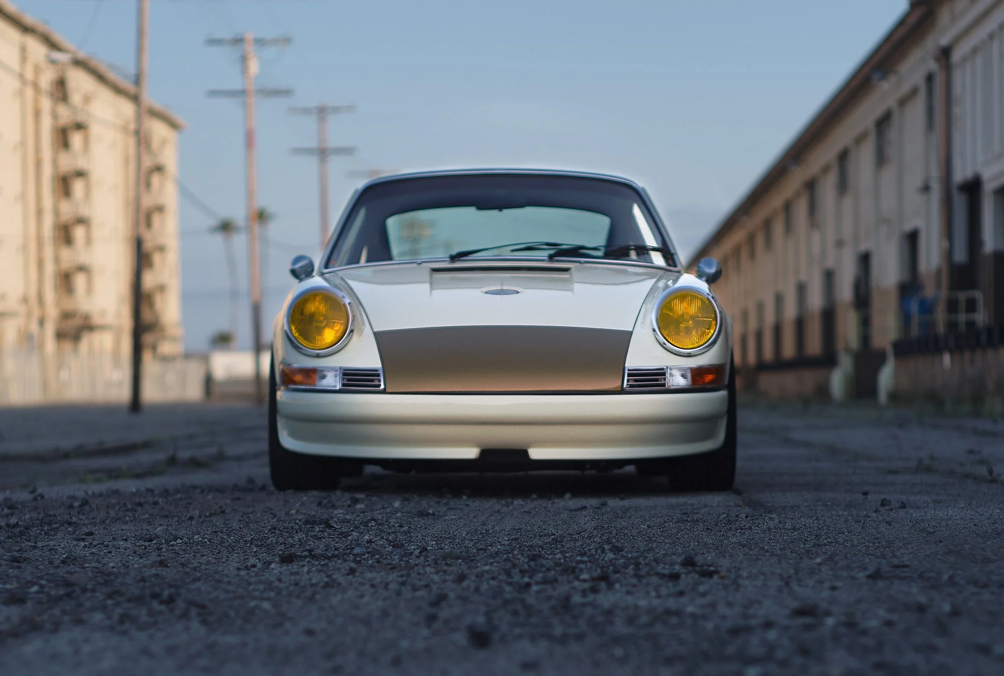 Front view of a vintage white sports car with yellow headlights on a gritty urban street between old multi-story buildings