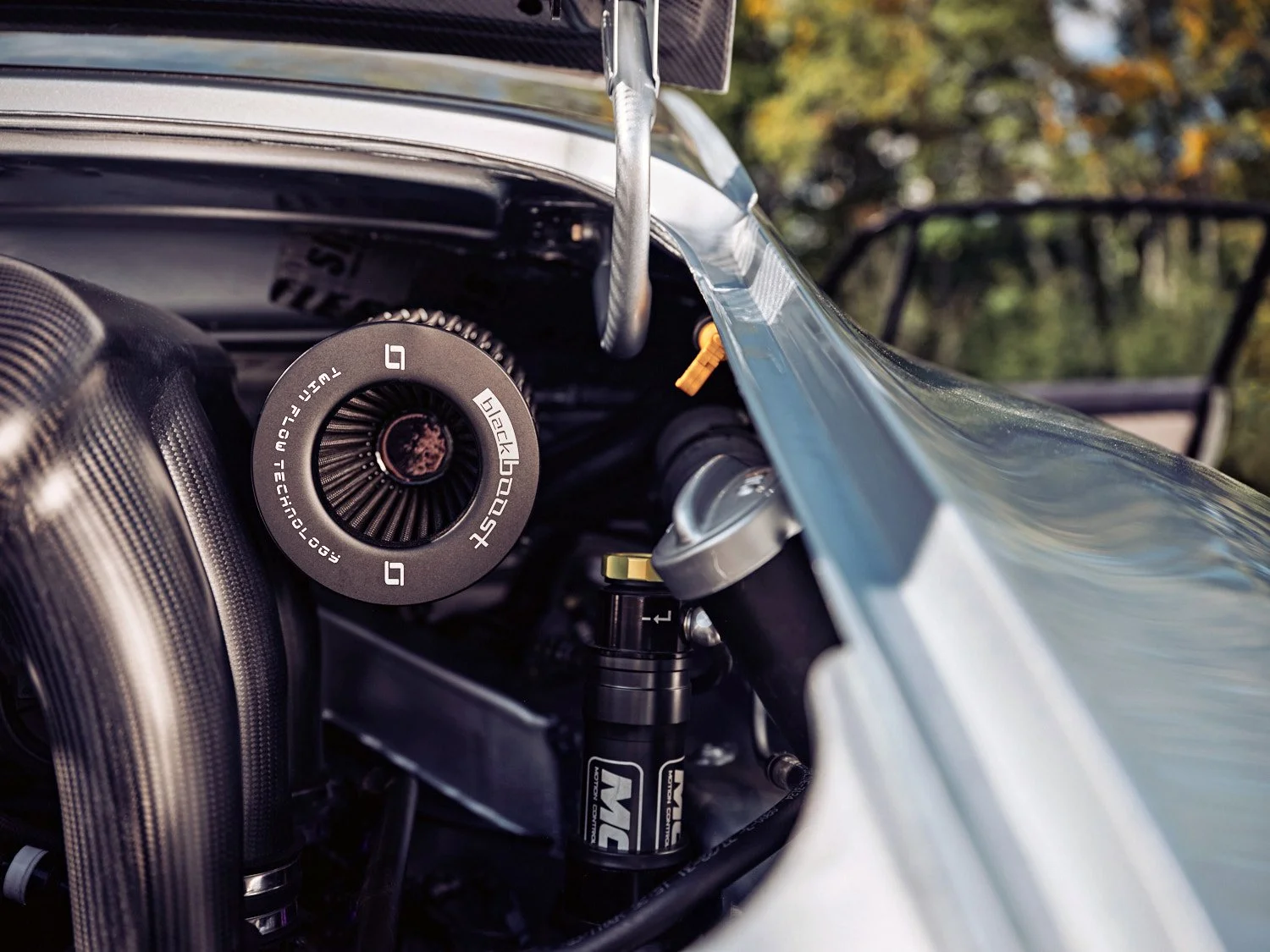 Close-up of a section of a race car's interior showing an air intake duct, a shock absorber with 'MC' branding, and part of the car's body with trees and sky in the background.