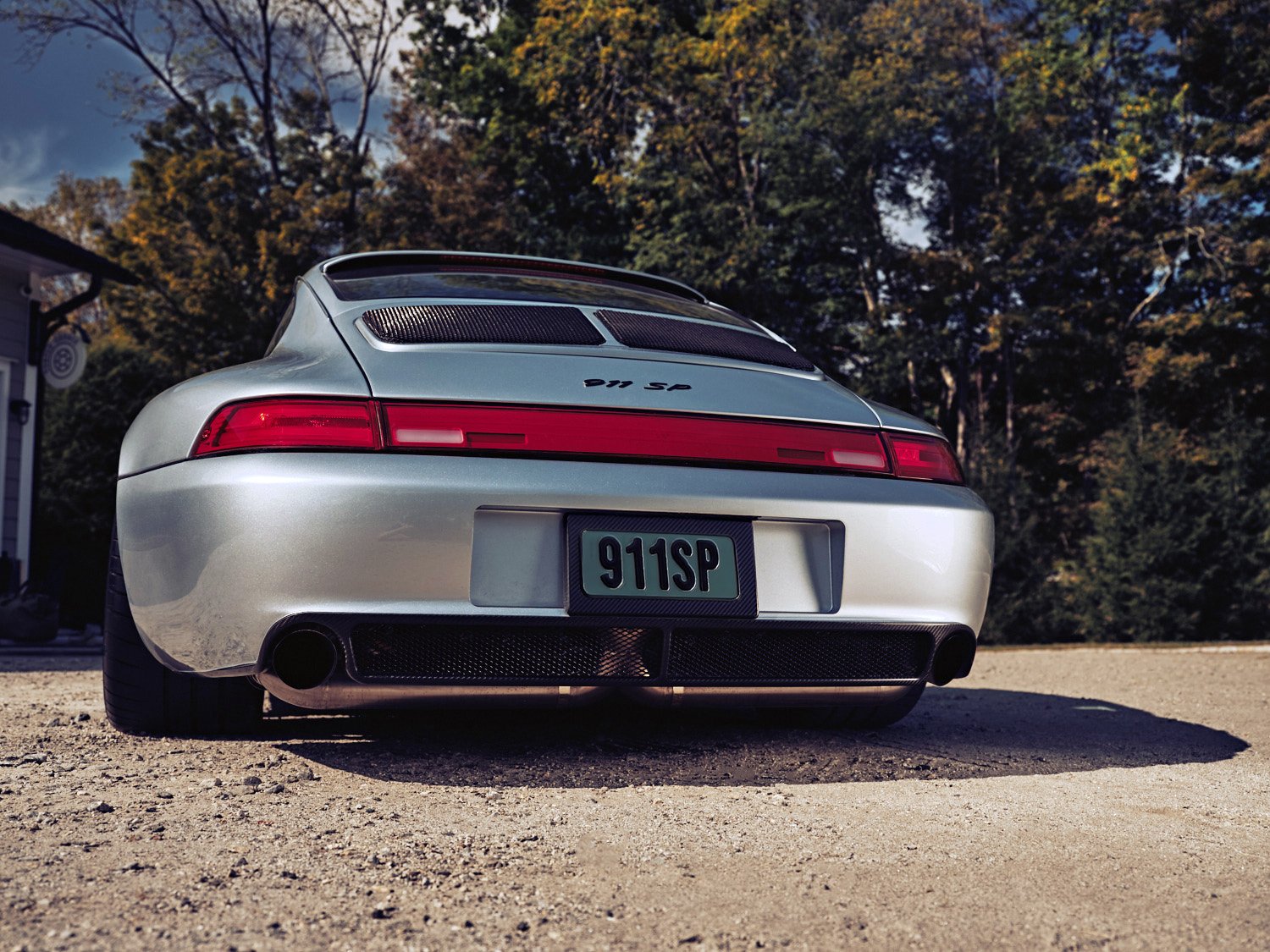 Rear view of a silver Porsche 911 SP parked on a dirt driveway, with a background of trees and a house, during daytime.