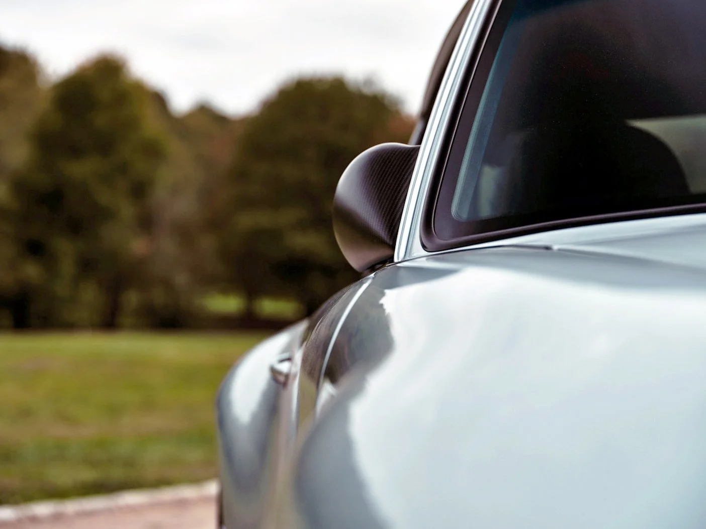 Close-up of the side mirror and part of the windshield of a silver sports car parked outdoors with trees in the background.