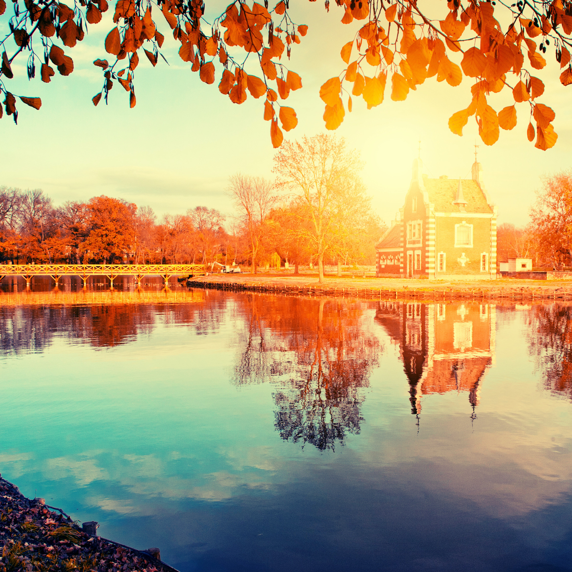 Autumn scene with a house near a lake, surrounded by orange and red trees, with their reflection visible in the water, and a bridge over the lake.