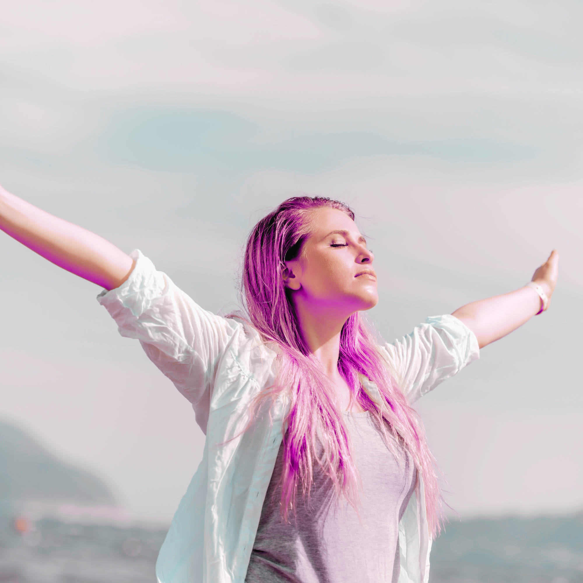 Woman with pink hair standing outdoors with arms outstretched, eyes closed, face peaceful.
