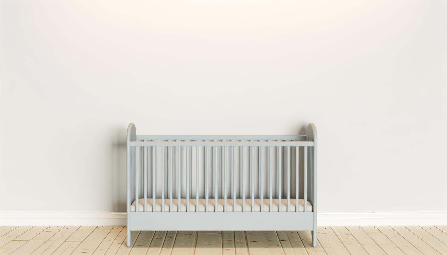 A white crib with a mattress in a minimalist room with wooden floor and a plain white wall.