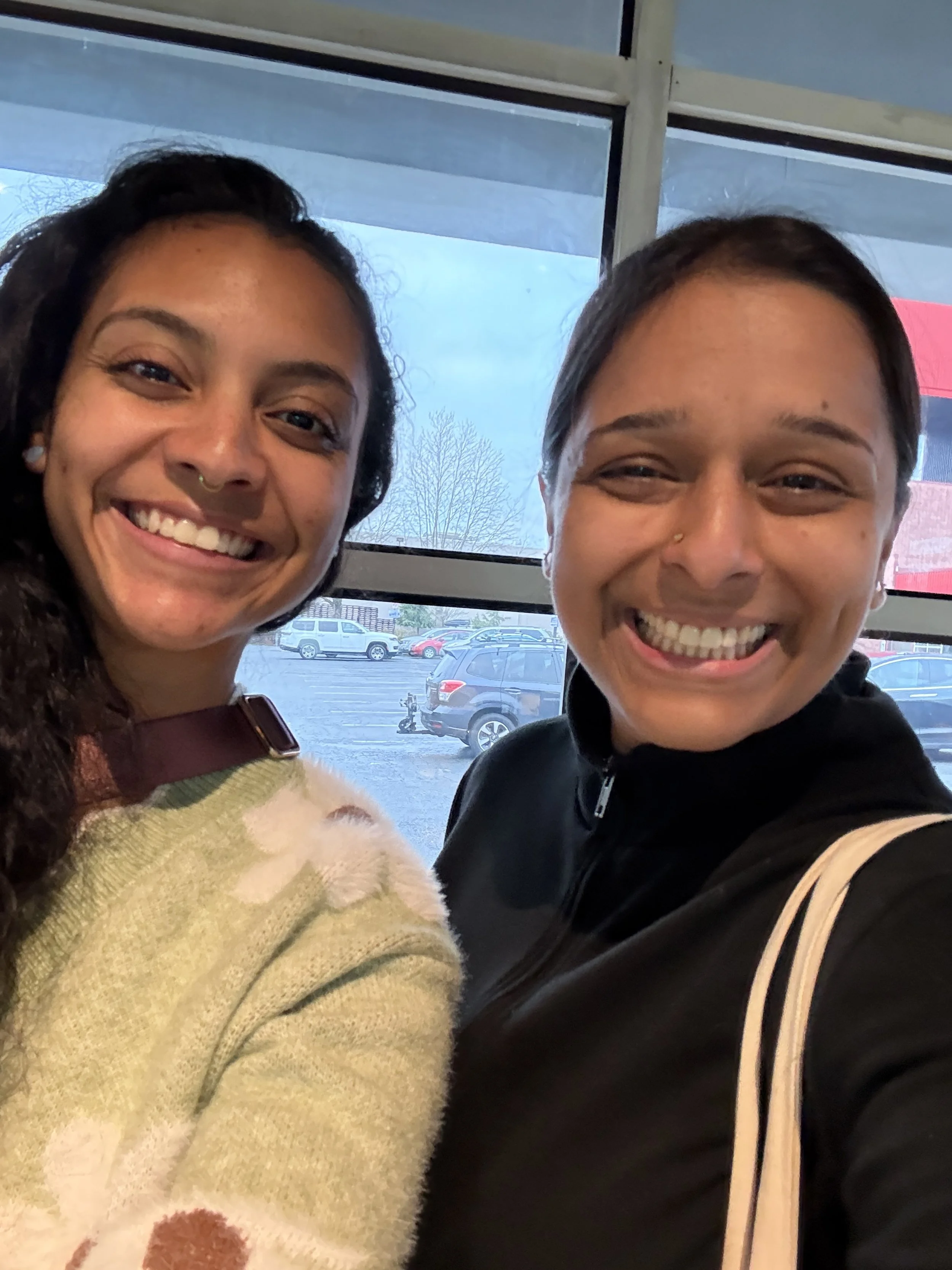 Two women smiling for a selfie in a parking lot near a large window with trees and parked cars visible outside.