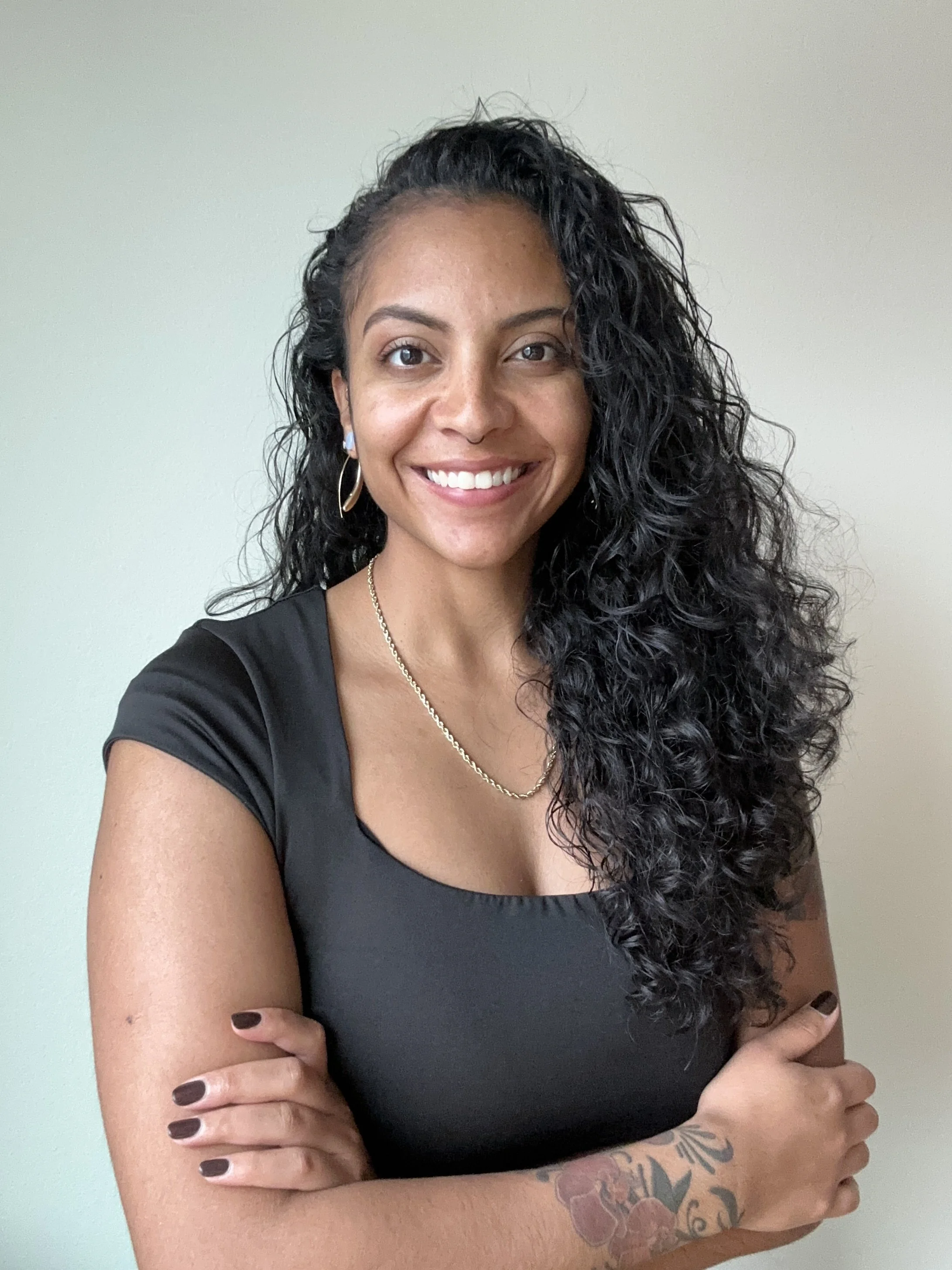 Kiona, a female presenting person with long, curly black hair, smiling, wearing a black top, gold hoop earrings, and a gold chain necklace, and sporting a tattoo on her forearm.