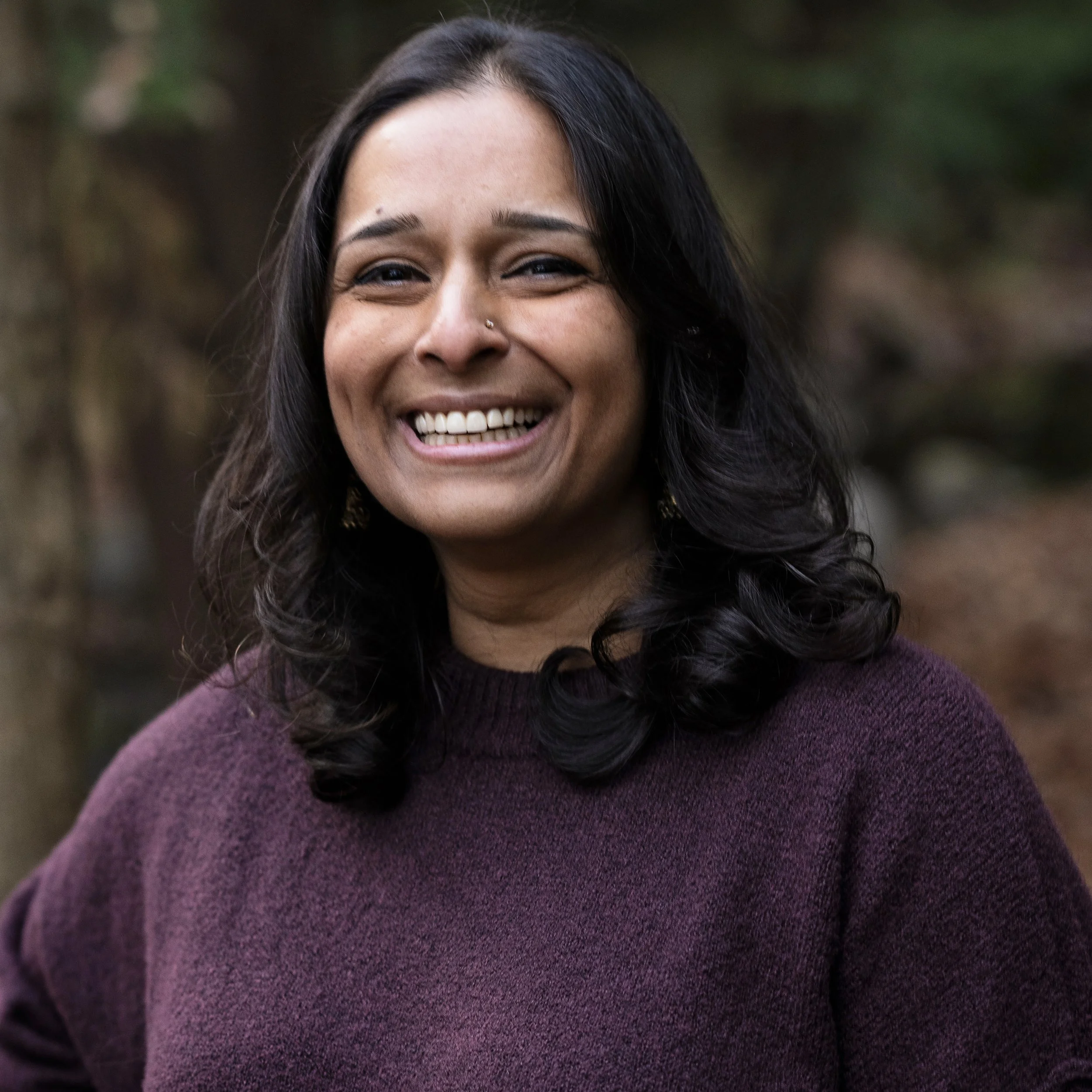 Jaya, a female presenting person with dark, wavy hair, smiling outdoors, wearing a purple sweater in a natural, wooded setting.