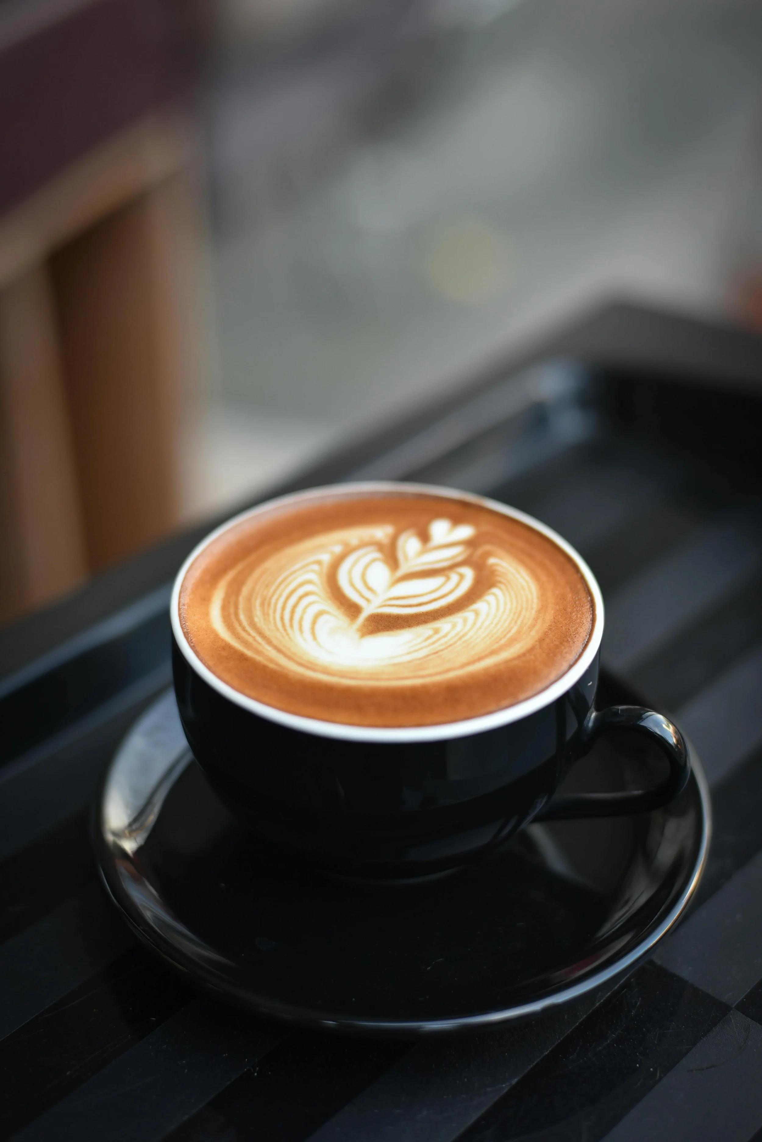 A black cup of coffee with latte art sitting on a matching black saucer on a dark wooden table.