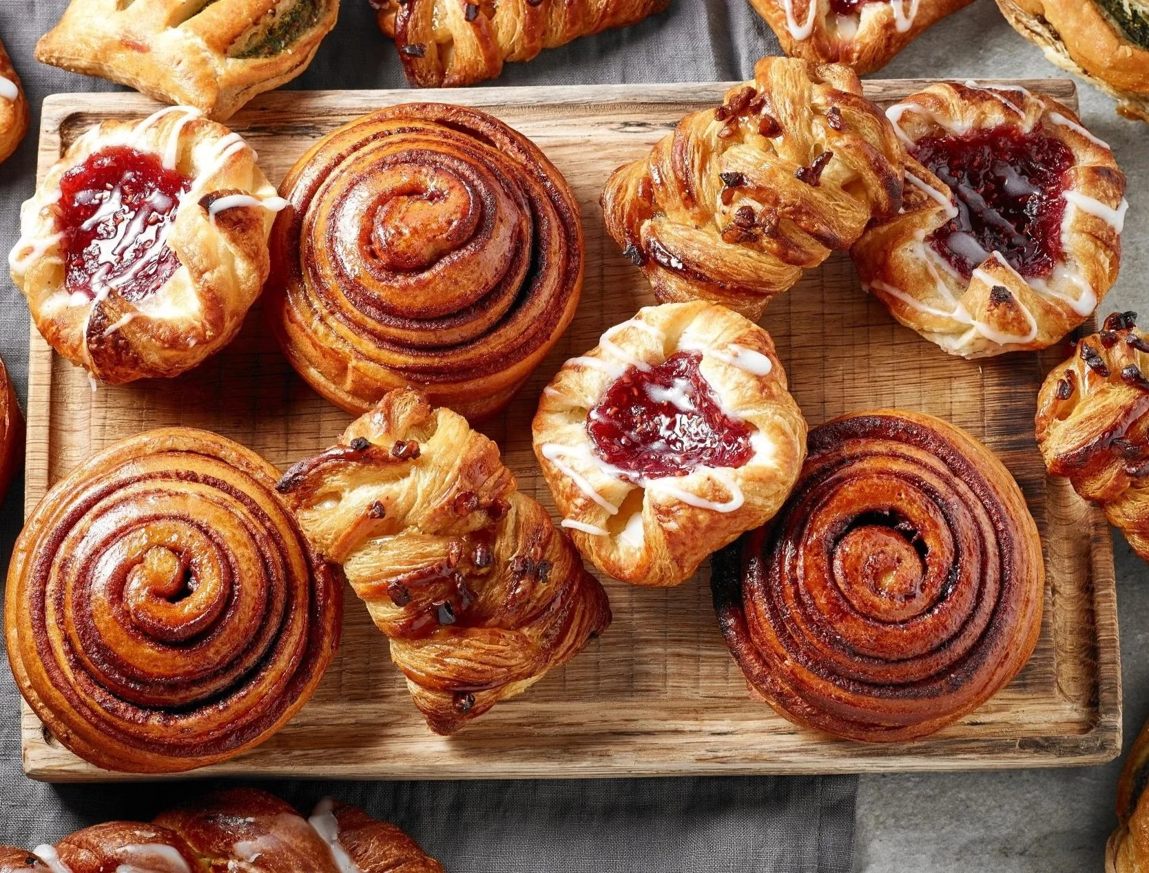 Assorted cinnamon rolls and Danish pastries on a wooden tray, glazed with icing and fruit jam.