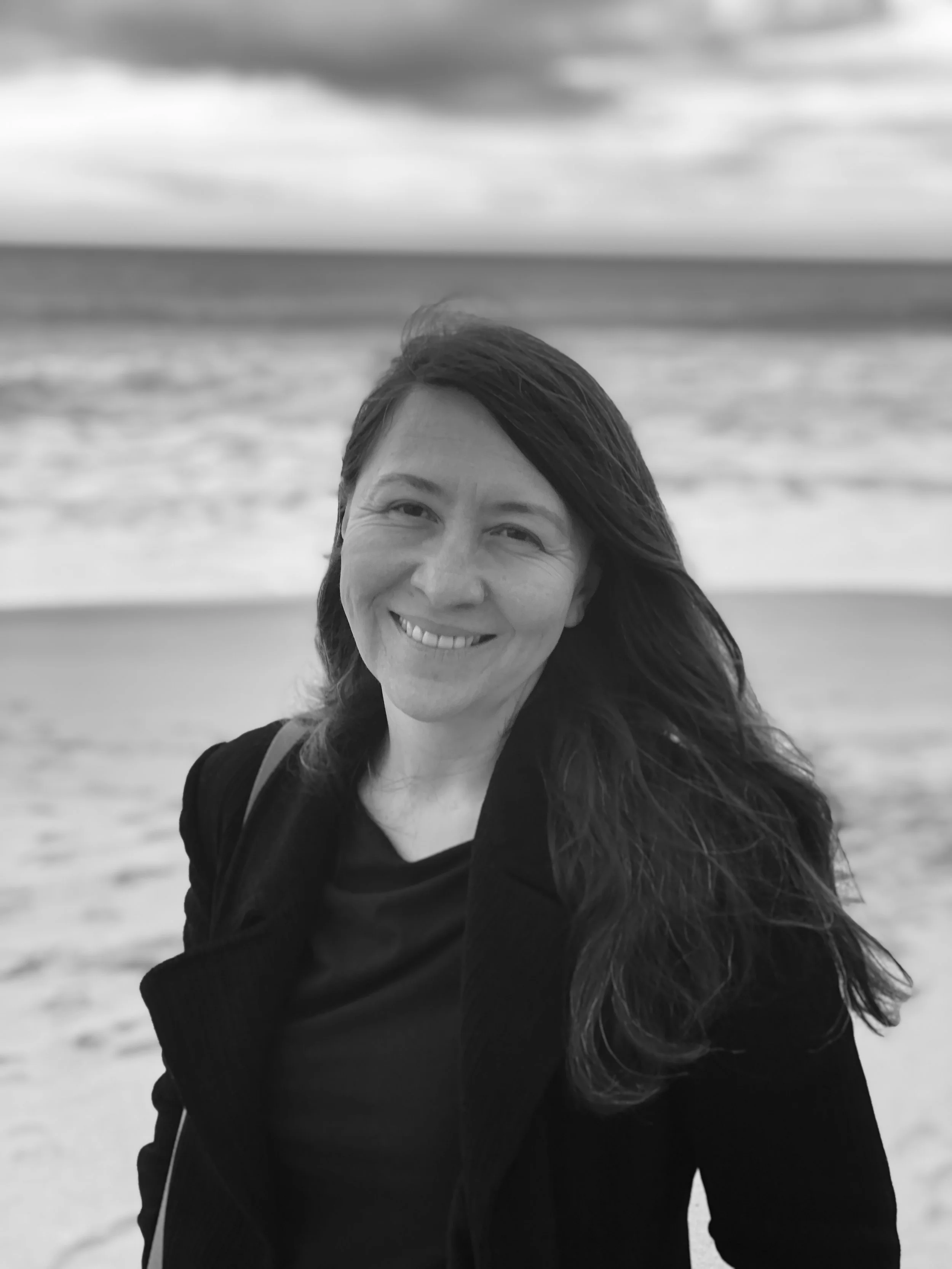 A woman smiling on a beach with waves and cloudy sky in the background, black and white photo.