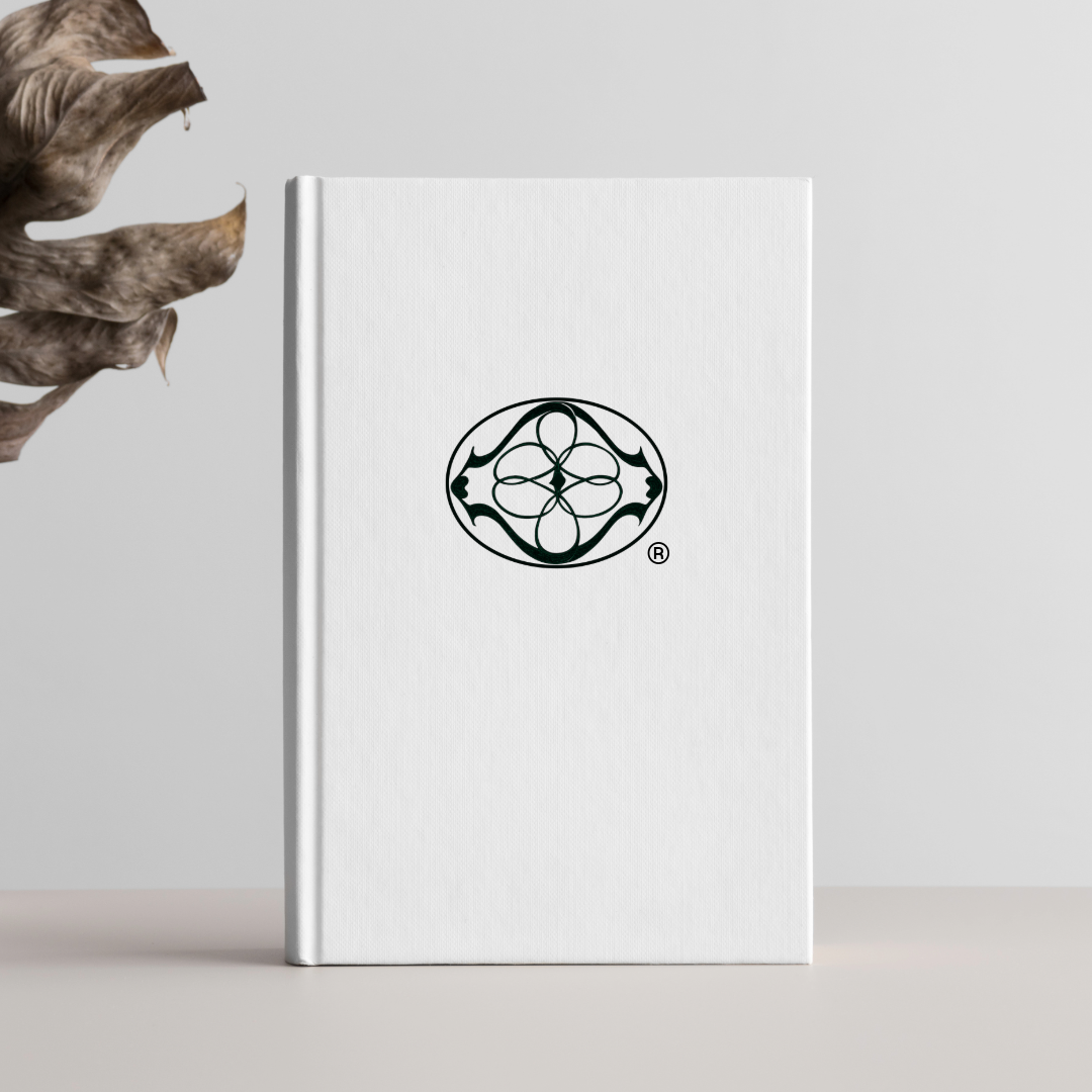 A white hardcover book with a black decorative logo on the front cover, placed against a plain white background, with dried leaves on the left side.