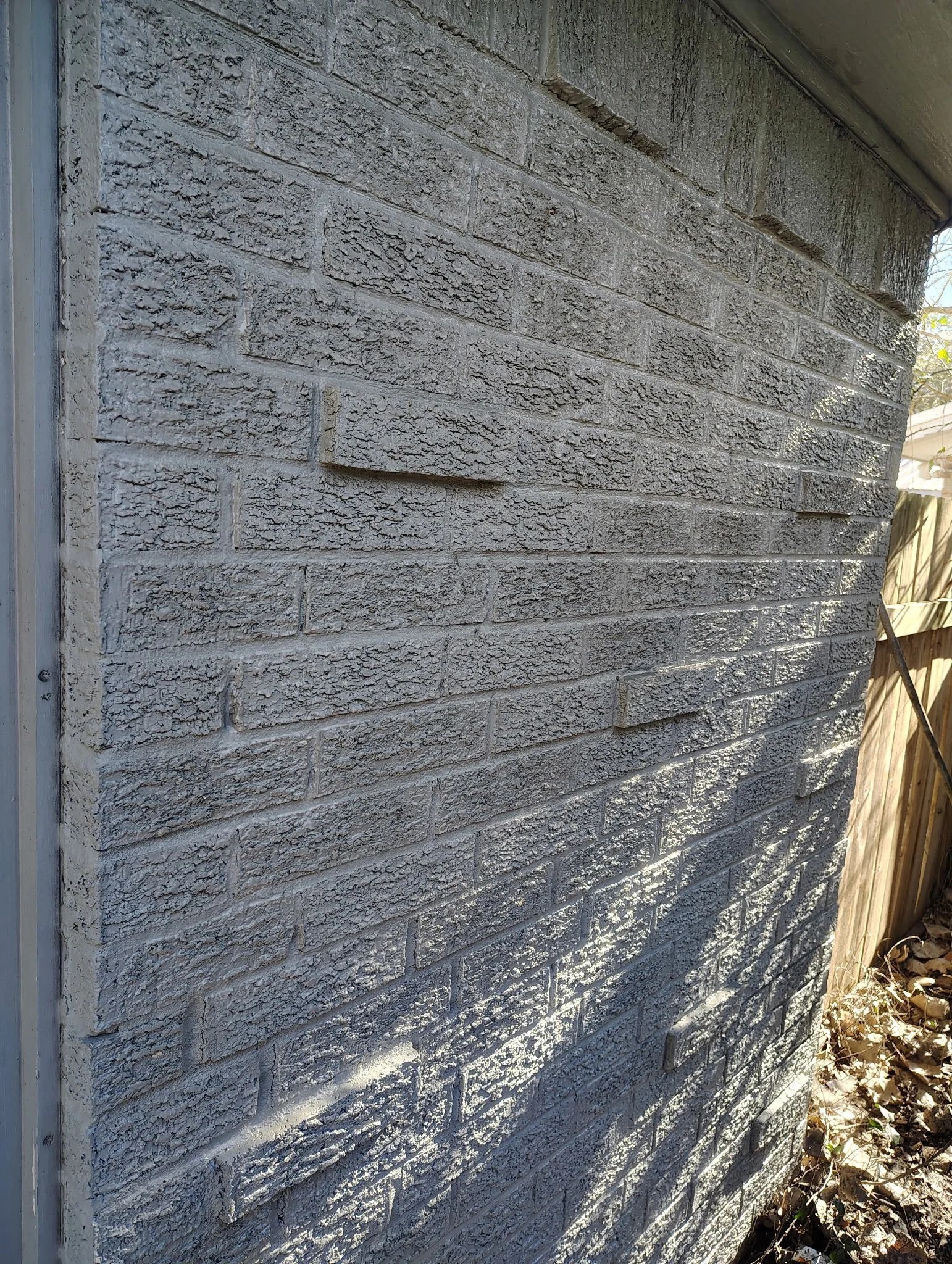 Close-up of a gray brick exterior wall with light shadow and a wooden fence on the right side.