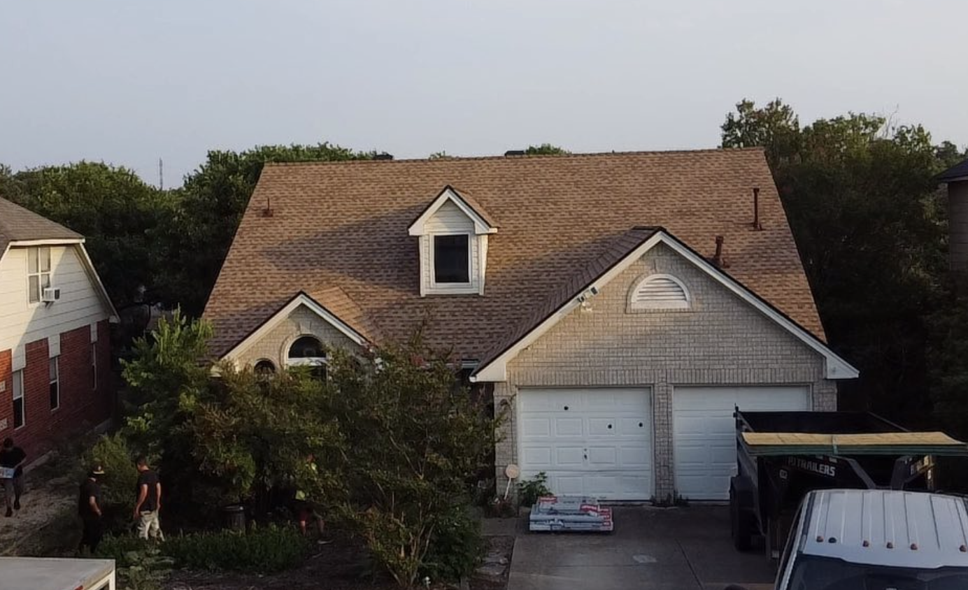 A two-story house with a brown roof, white brick facade, and attached garage, surrounded by trees and neighboring houses, with people working outside.