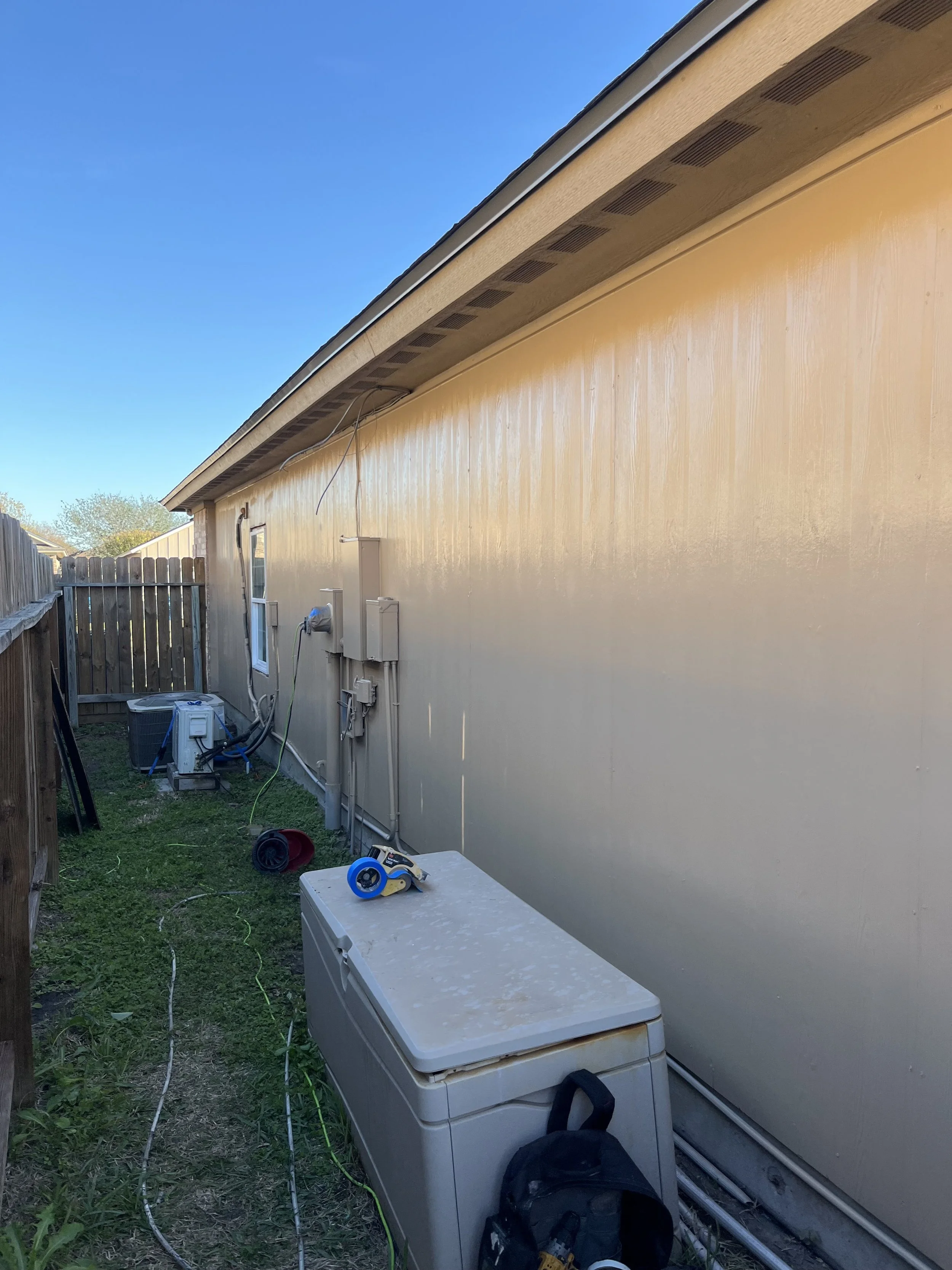 Side of a house with a beige wall, window, fenced backyard, and various utility and cleaning equipment, including a cooler, backpack, and tape measure, under a clear blue sky.