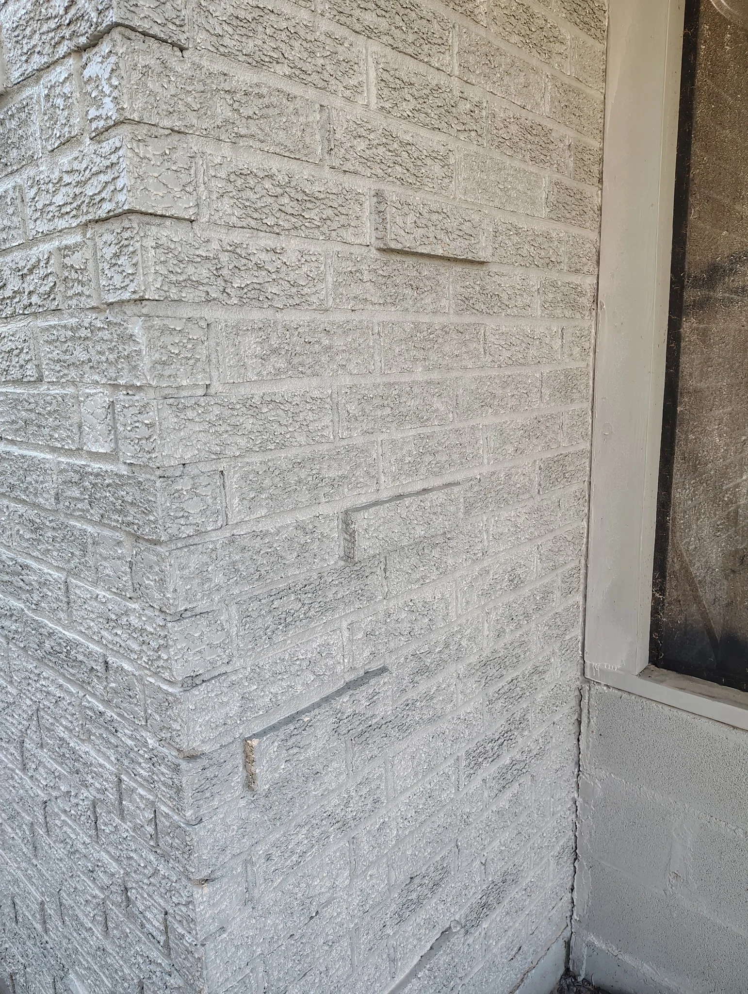 Corner of a beige brick wall next to a window with a white frame and painted white brick wall below.