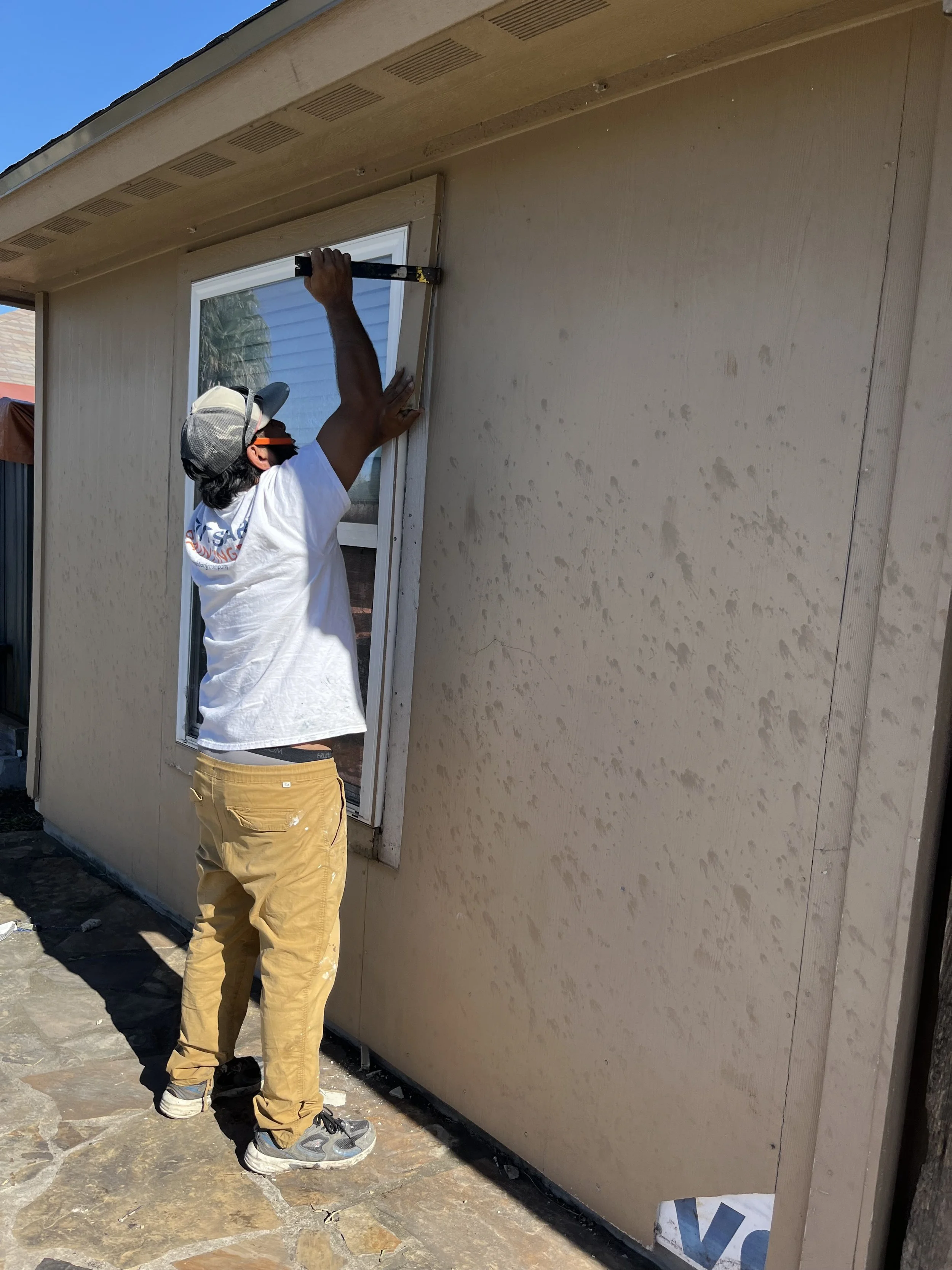 A man installing or repairing a window on the exterior of a house. He is wearing a hat, sunglasses, a white t-shirt, tan pants, and sneakers, and is using a level tool.