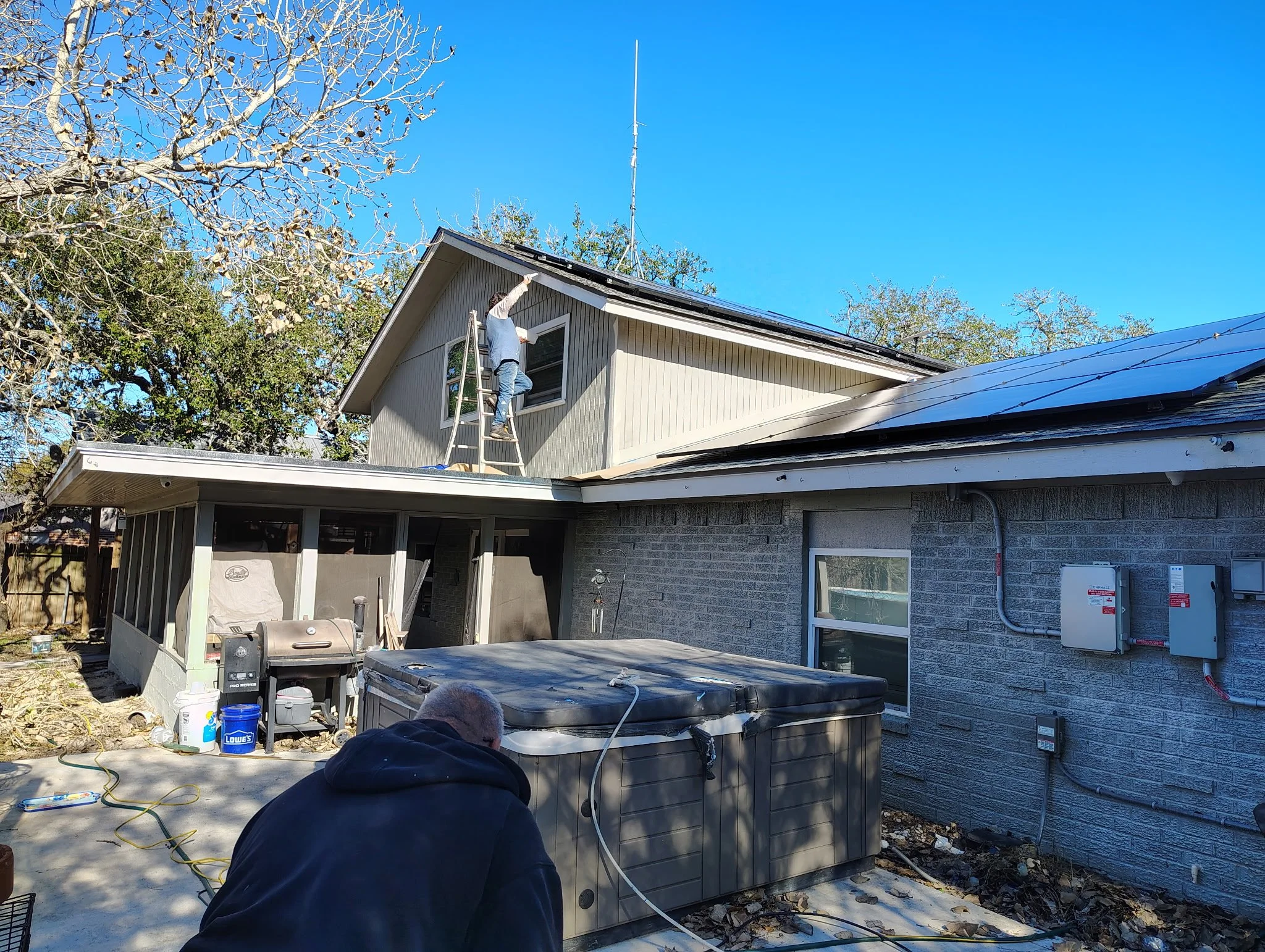 Two people working on a house exterior. One person is on a ladder working on a window, while the other is kneeling on the ground nearby. The house has a screened porch, gray brick siding, and solar panels on the roof. There are trees in the backgroun