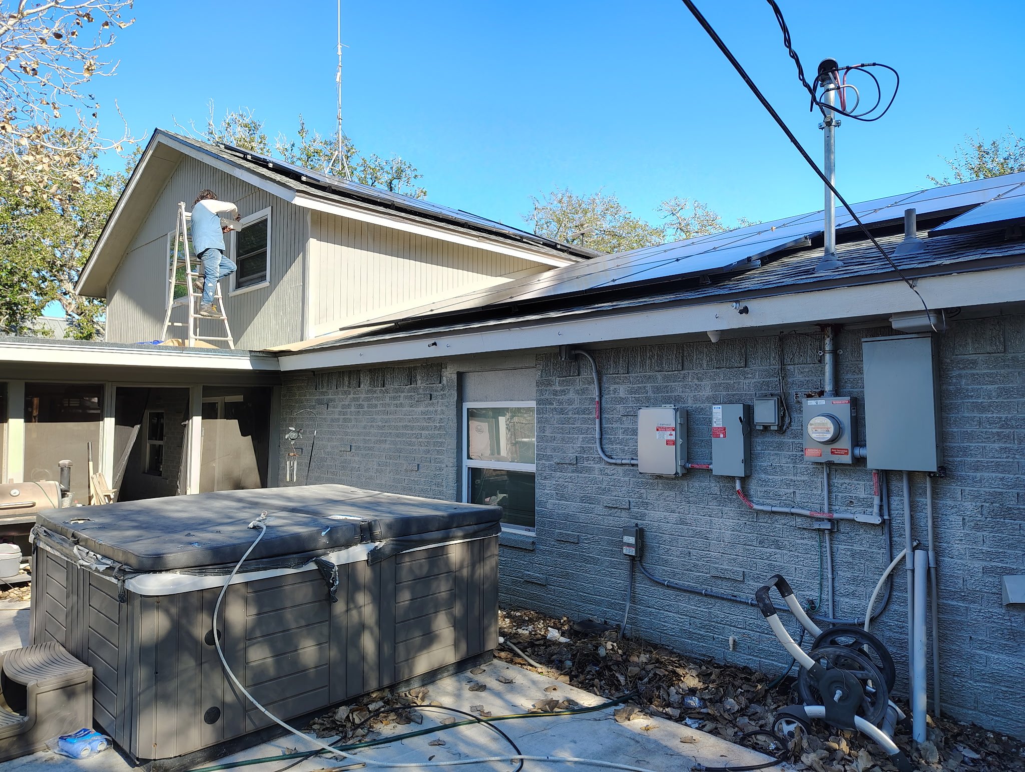 A person standing on a ladder working near a window on the second floor of a house, with electrical and utility boxes on the brick exterior wall, a hot tub in the backyard, and a clear blue sky.