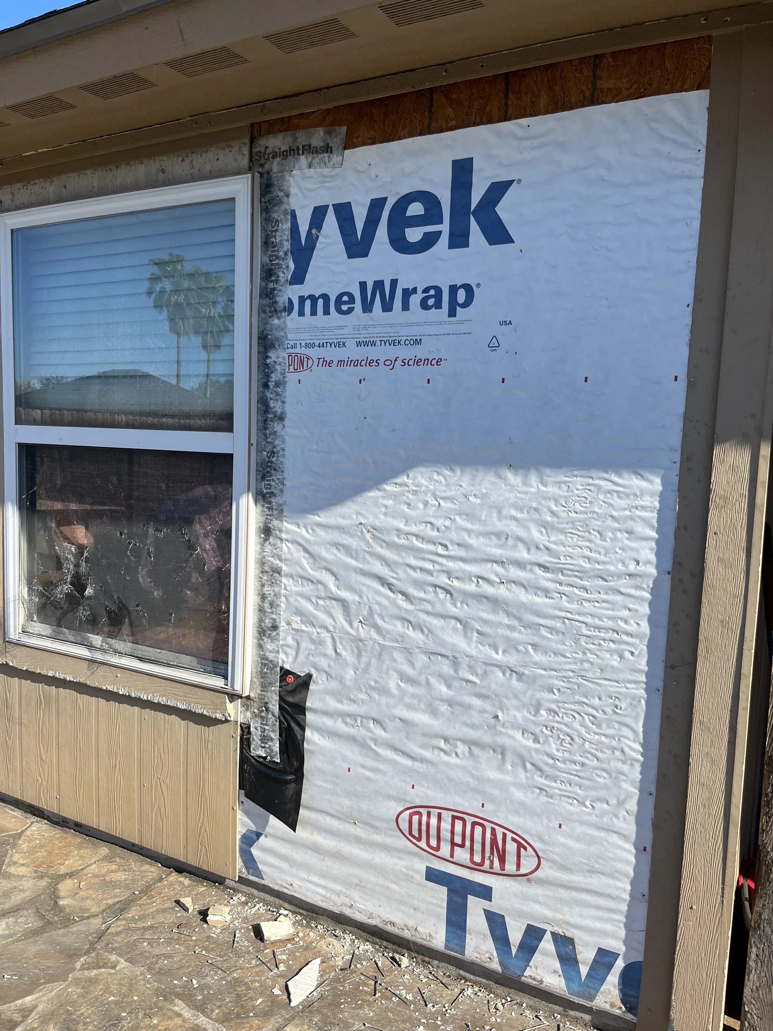 Building exterior with wall insulation material partially installed near window, with construction debris on stone-paved ground.