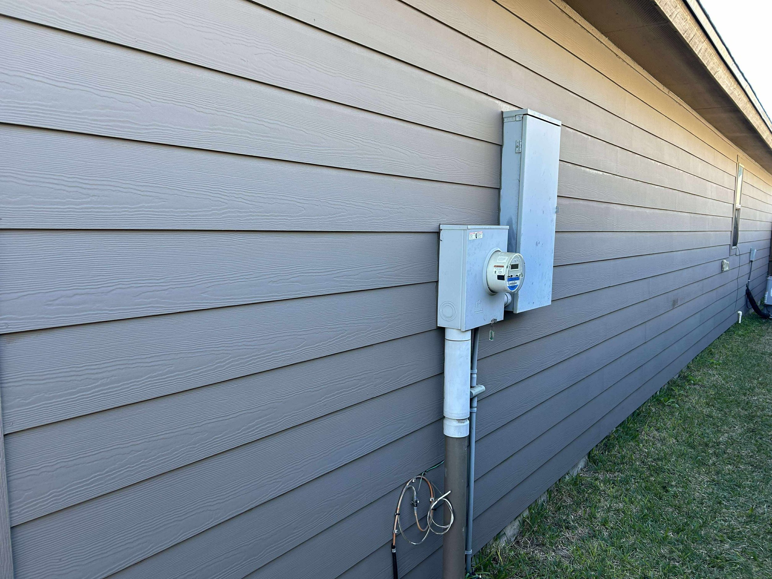 Side of a house with horizontal siding, electrical meter, and utility boxes attached to the wall, with some grass at the bottom.