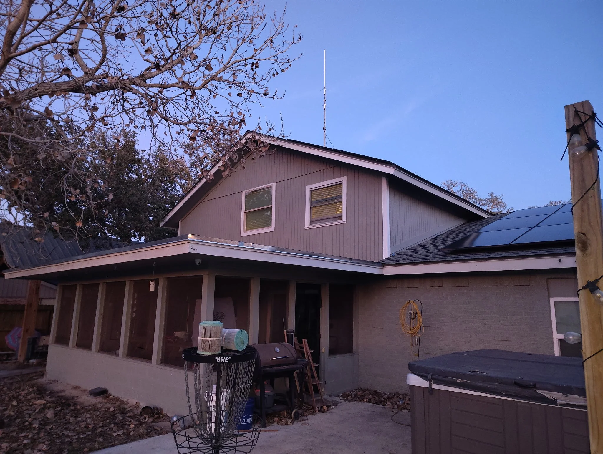 Backyard of a house at dusk, showing a screened porch, a hot tub, a basketball hoop with a basketball, and a leafless tree.
