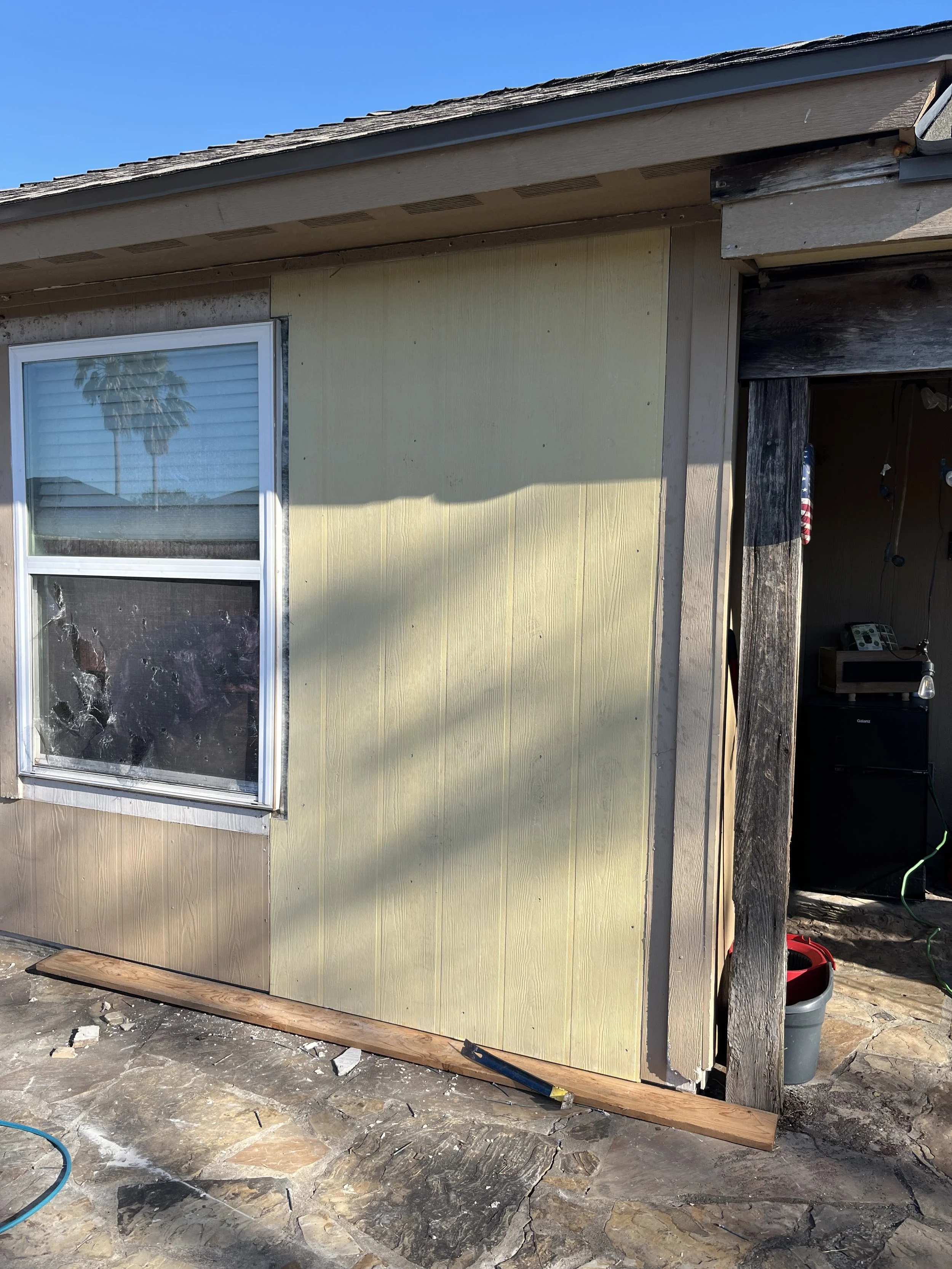 Partially renovated exterior of a house with beige and yellow siding, a window with blinds, and a damaged corner with exposed wood and construction tools.