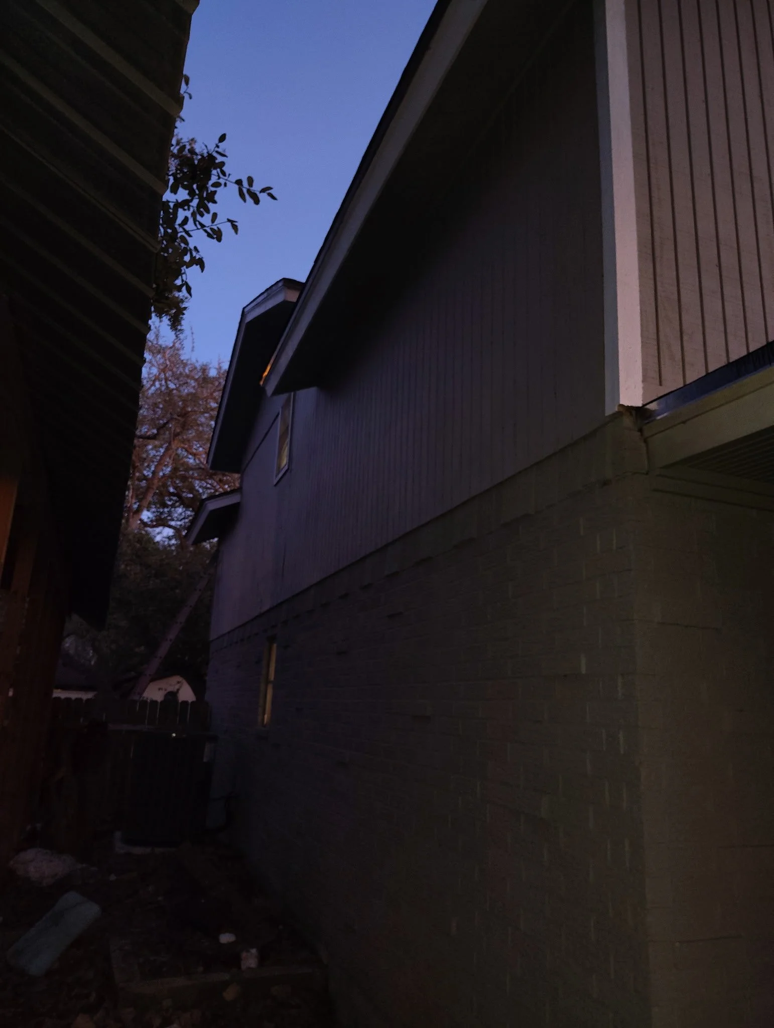 Photo taken during twilight showing side view of a house with siding and brick wall, with a small window and trees in the background.