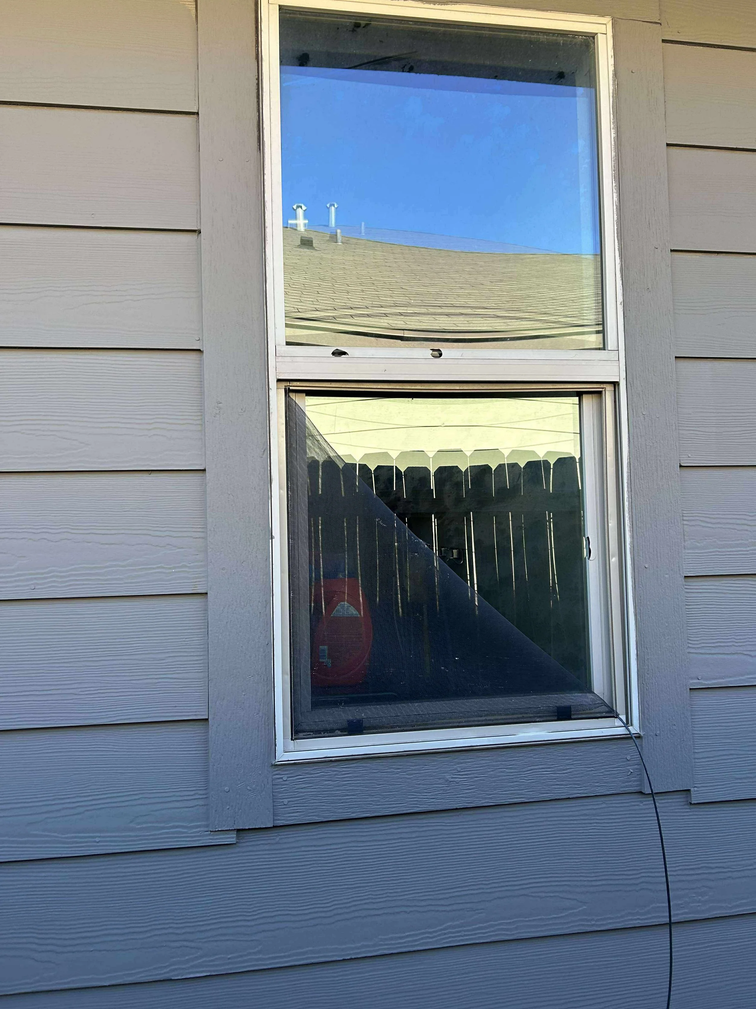 A house with gray horizontal siding, showing two rectangular windows. The top window reflects a blue sky and roof, while the bottom window shows a black fence and a red garden tool inside the house.