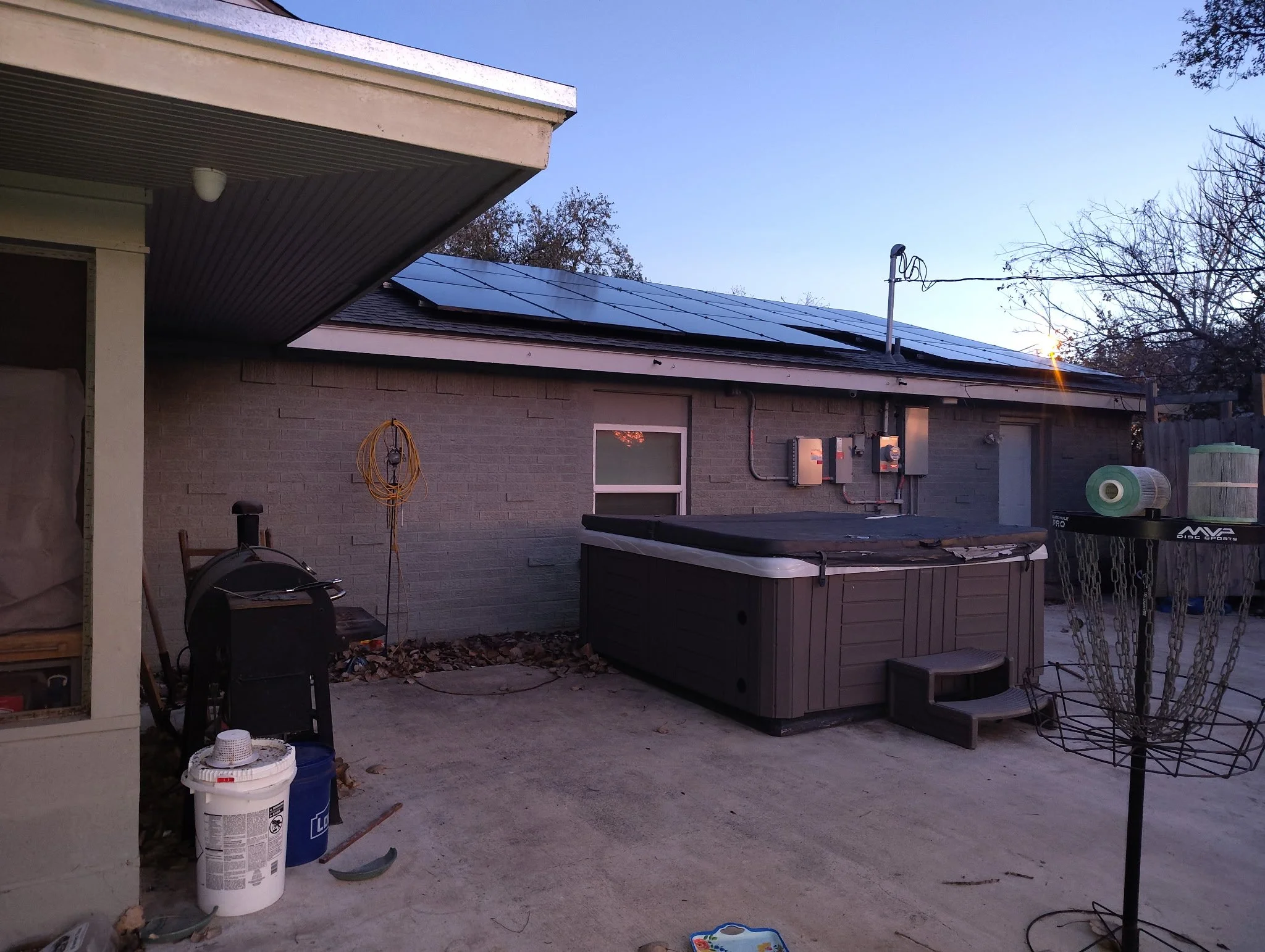 Backyard patio with a hot tub, an equipment stand with a disc golf basket, and solar panels on the roof of a house during dusk.