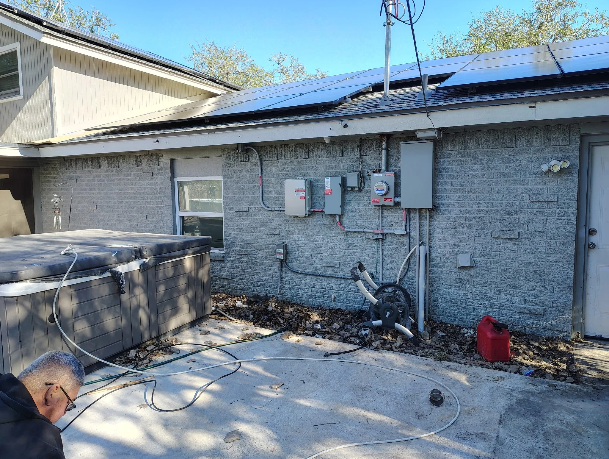 Backyard with a gray brick house wall, showing various electrical panels and meters, an above-ground hot tub, vacuum hose, and a red fuel container on the ground. A man with glasses in the lower left corner is working.