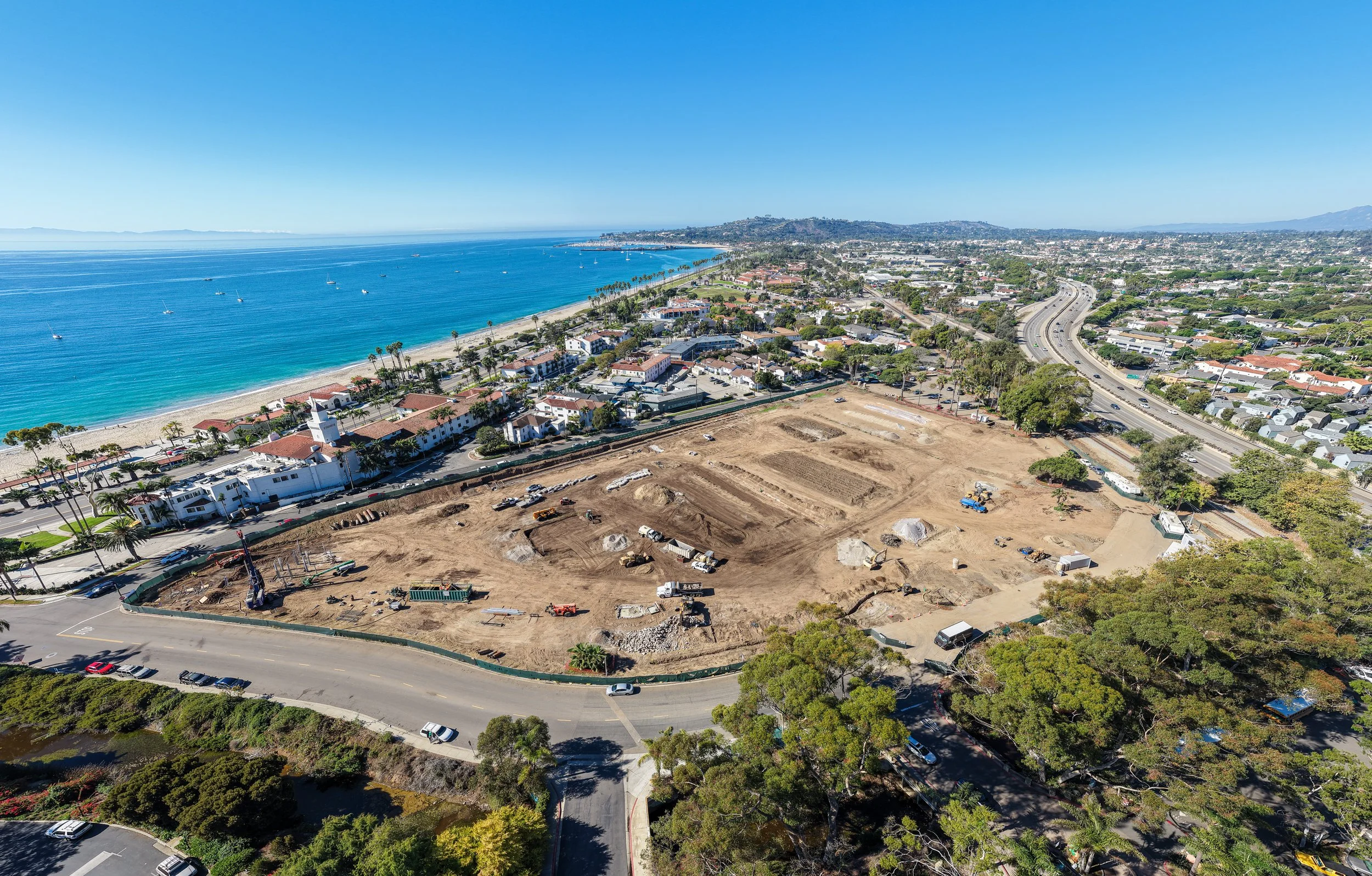 Aerial view of a construction site near a beach with several buildings and a highway in the background.