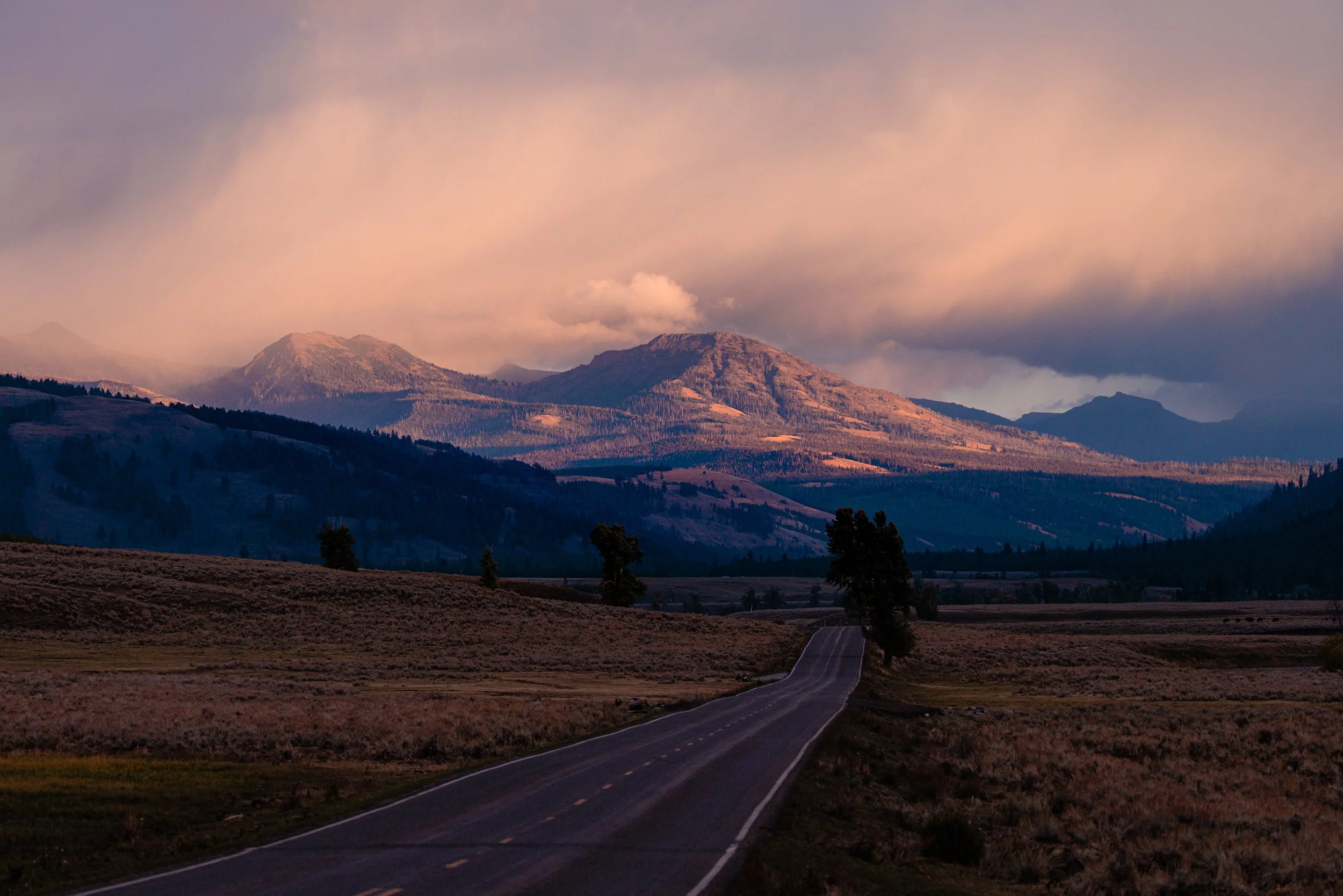 A long open road leading through a grassy plain towards distant mountains with colorful clouds overhead at sunset.