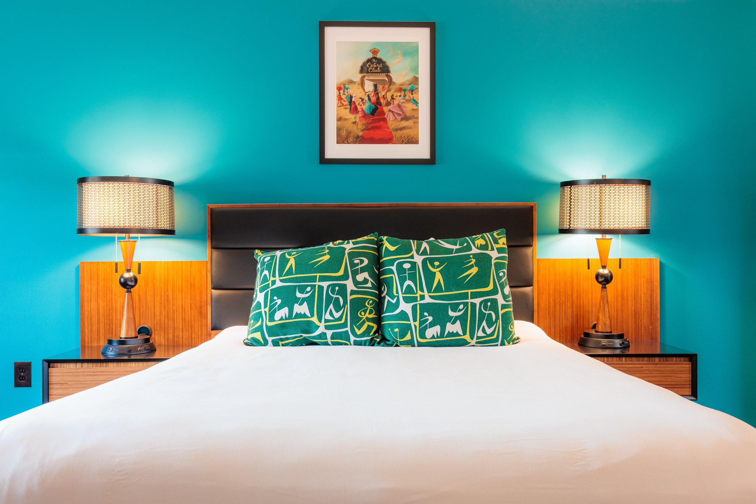A neatly made bed with two green patterned pillows in a modern bedroom featuring teal walls, wooden side tables with matching lamps, and a framed colorful artwork hanging above the headboard.