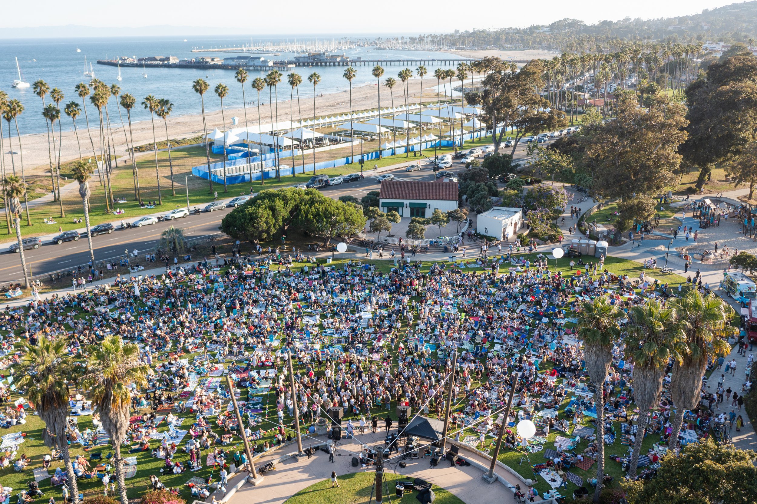Crowd gathered on a grassy area near a beach with palm trees, tents, and a stage, with the ocean and sailboats in the background.