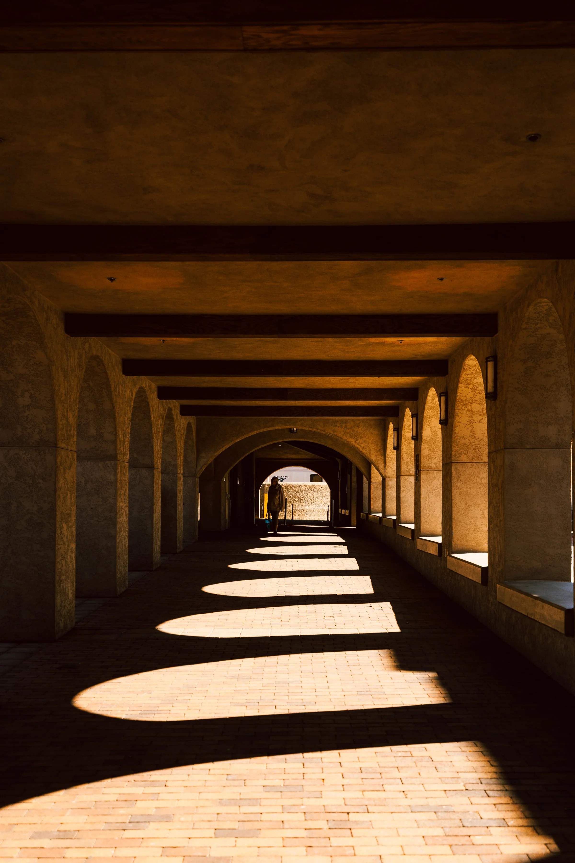 A person walking through a covered walkway with arched openings on the right side, casting shadows on the brick floor, with sunlight at the end of the corridor.