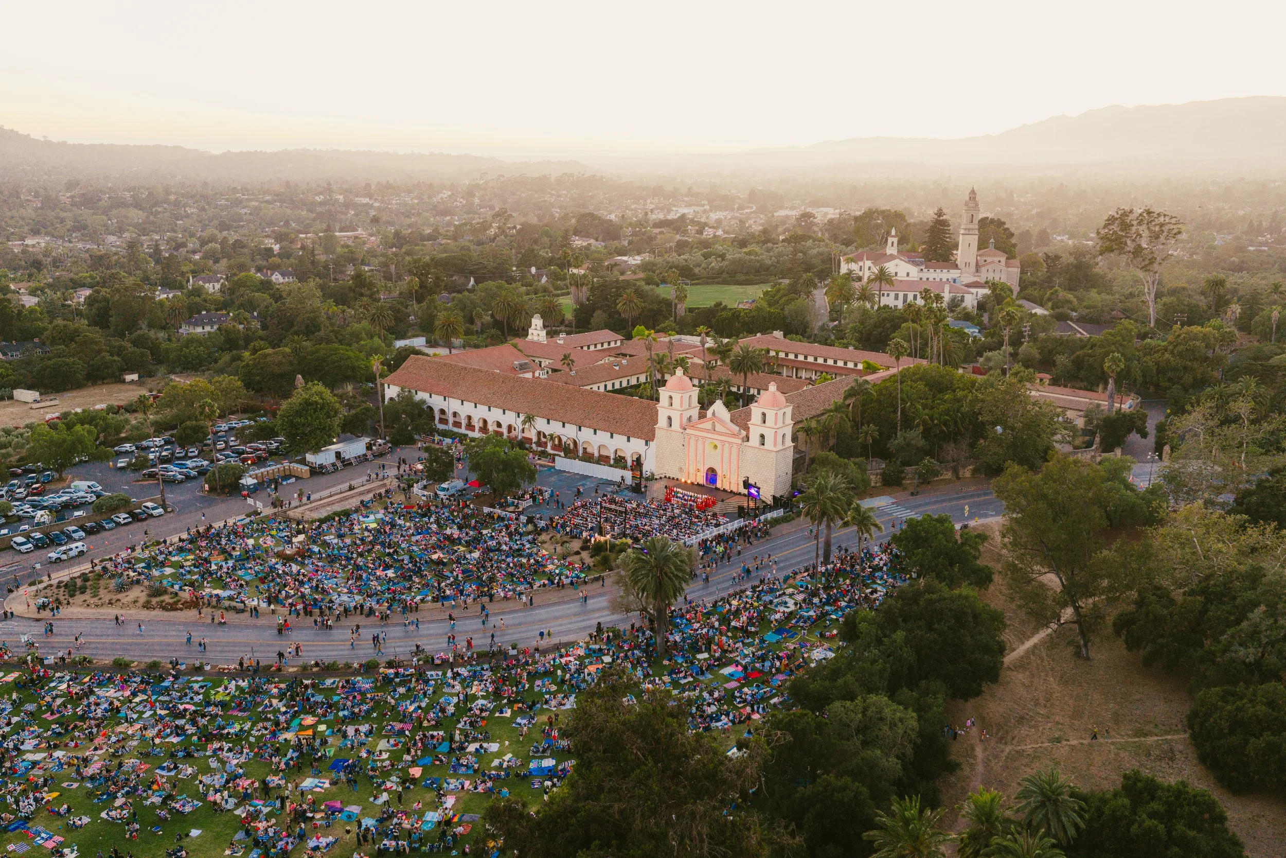 Aerial view of a large outdoor gathering with people, umbrellas, and chairs in front of a church and historic buildings in a lush, green area at sunset.