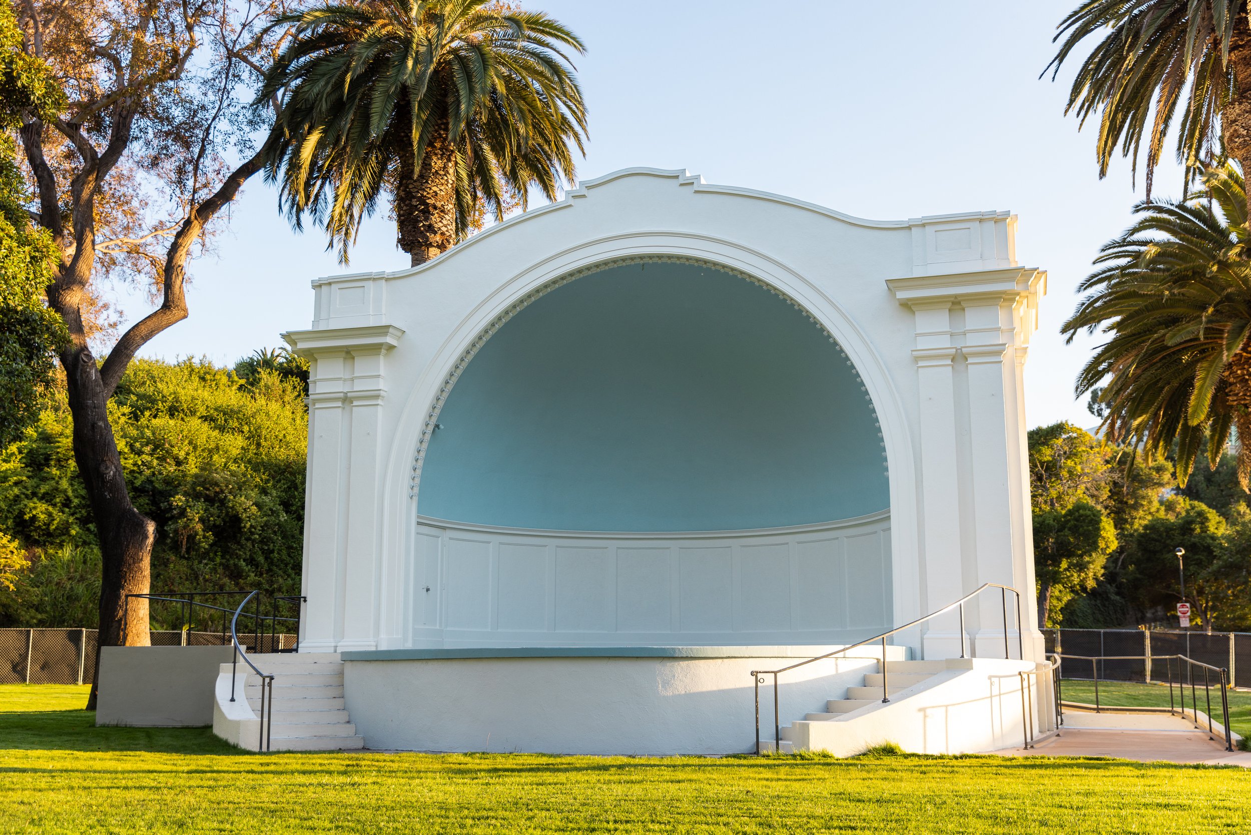 An empty outdoor stage with a white structure and a blue ceiling, surrounded by trees and grass